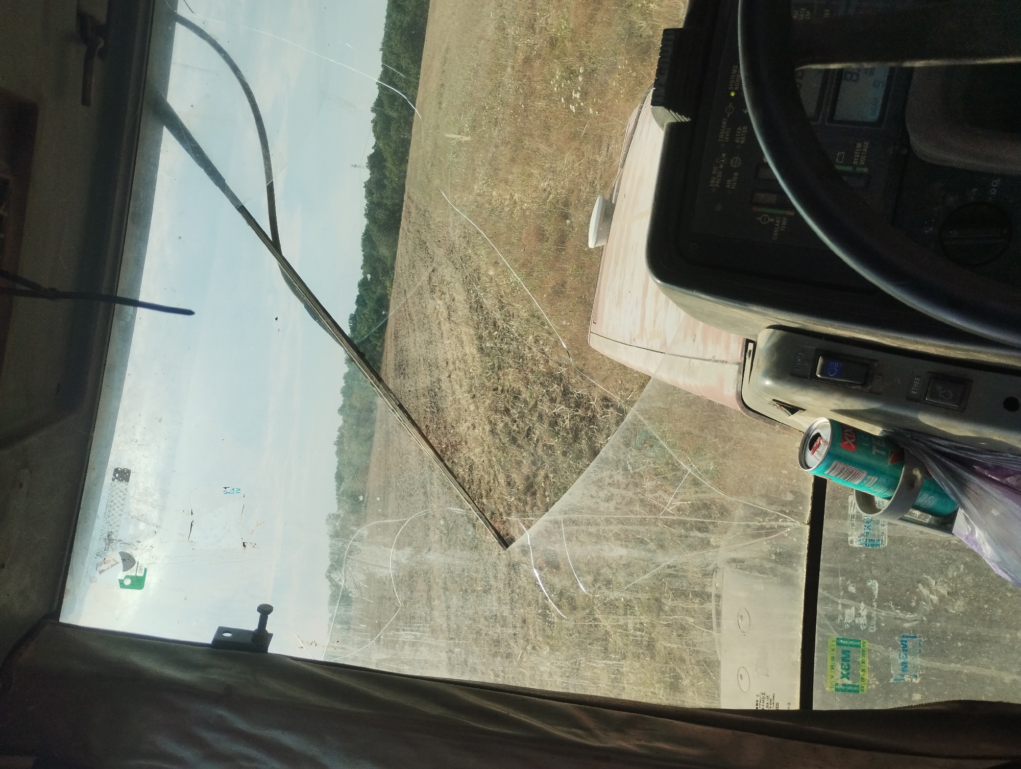 This is a first-person perspective image taken from inside a tractor cab, looking out through a cracked windshield. The scene depicts a rural environment, likely a field that has recently been worked, with dry, stubbled crops. In the background, there is a dense line of trees against a bright, hazy sky.
The interior of the cab is visible, showing part of the dashboard with various gauges and a steering wheel. A can of what appears to be a beverage is secured in a holder to the right of the driver. A brown curtain hangs on the left side of the windshield. The windshield itself has several prominent cracks, indicating damage.
The location of Kubadin, Bulgaria, is stated in the prompt. Given the agricultural setting, the scene likely takes place on farmland surrounding the village. No people are visible in the image. The overall impression is of a working tractor operating in a rural agricultural area.