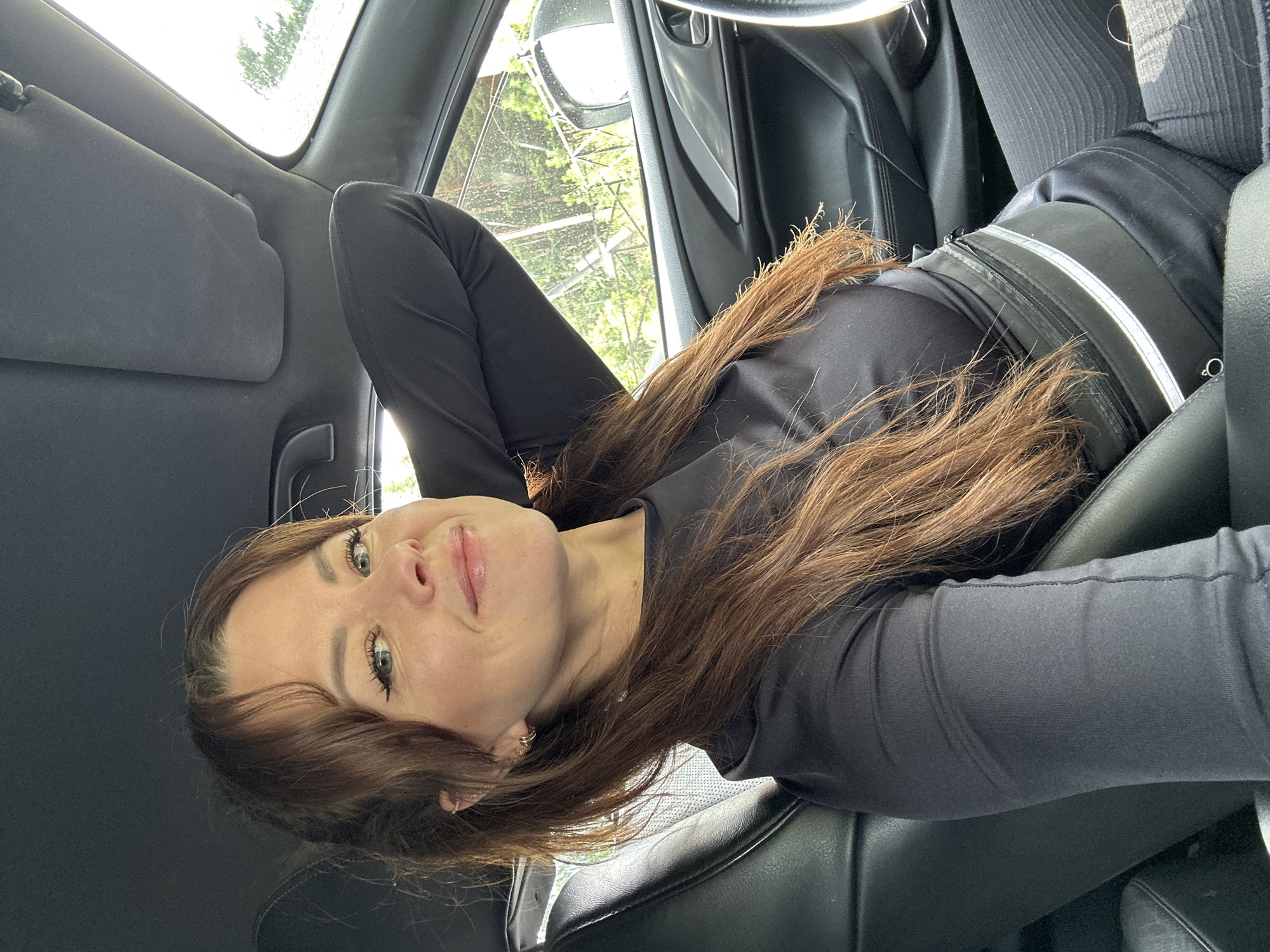 A woman with brown hair and blue eyes is sitting in the driver's seat of a car. She is wearing a black long-sleeved shirt and black pants. Her left arm is resting behind her head. She is smiling at the camera. The interior of the car is visible, including the leather seats, dashboard, and steering wheel. Outside the car, trees and a cloudy sky are visible through the windows. There is no information or visual cue in the image to indicate that this scene is located in Chaikovsky, Russia.