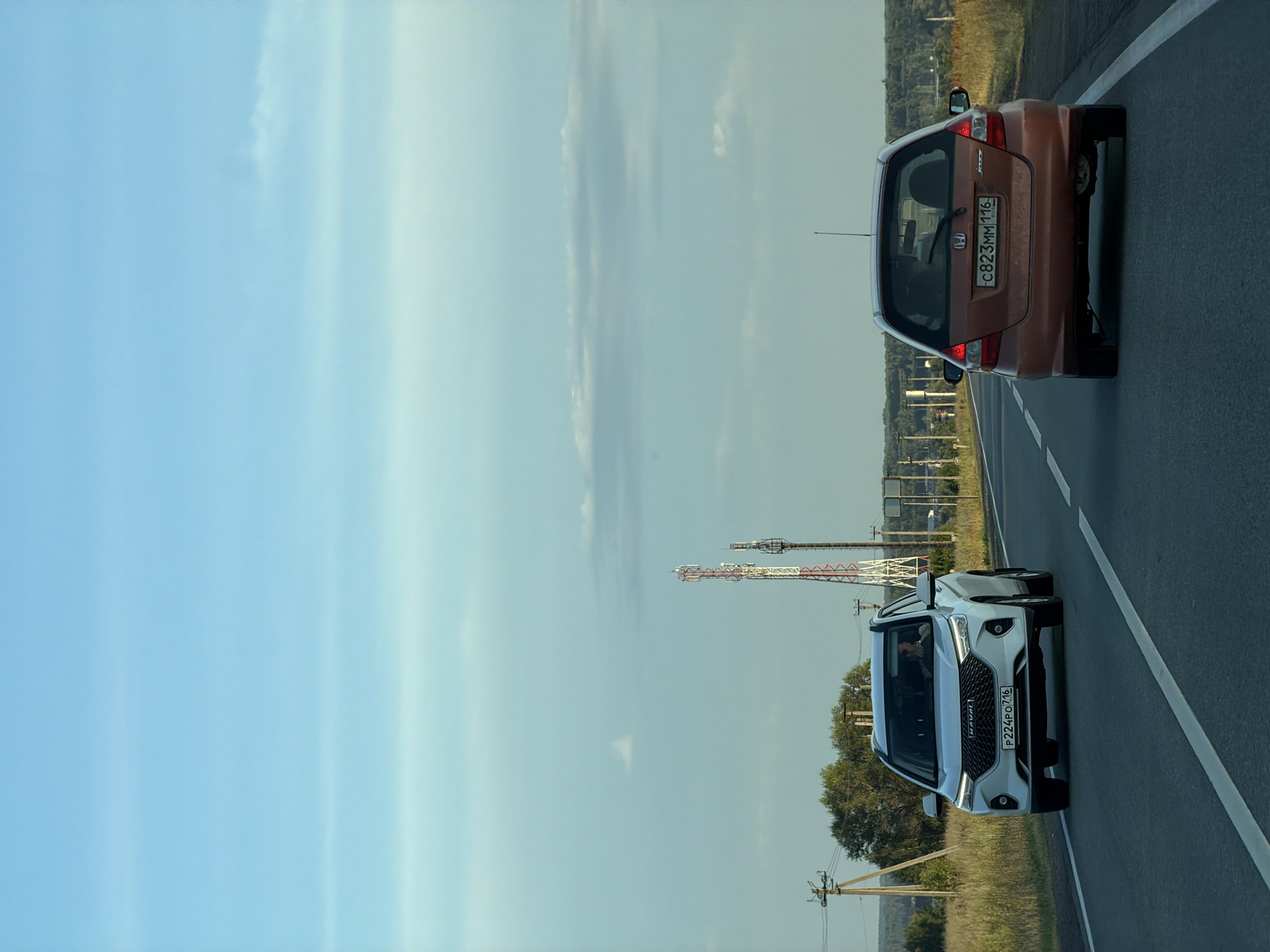 Two cars are traveling on a paved road during the daytime. The car in the foreground on the left is a white Haval SUV, facing forward. The car on the right is an orange Honda Jazz hatchback, also facing forward and slightly ahead of the Haval. The road has white lane markings. The environment is rural, with dry grass and trees on either side of the road. In the distance, there are power lines, communication towers, and a tree line. The sky is blue with some wispy clouds. The license plate of the Haval reads "P224PO 716". The license plate of the Honda Jazz reads "C823MM 116". The scene suggests a journey on a highway outside of a populated area, possibly in the vicinity of Обухово, Russia, given the presence of typical Russian license plate formats.