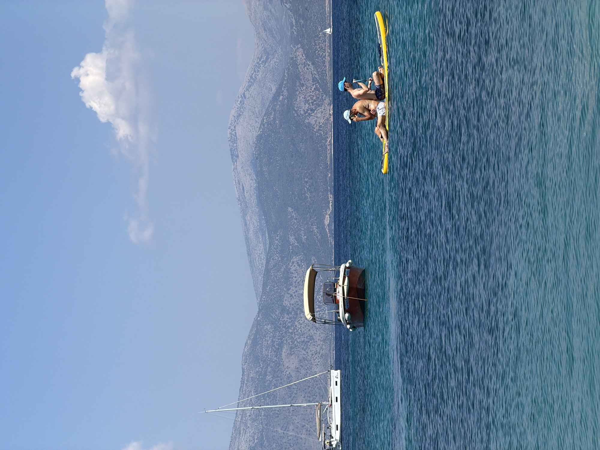 A wide shot captures a tranquil scene in the Municipal Unit of Kastos, Greece, under a clear blue sky with a few scattered clouds. In the foreground, the turquoise water is calm, with gentle ripples.
Dominating the middle ground are several watercraft. To the left, a white sailboat with its mast pointing skyward is anchored. To its right, a small, red motorboat with a tan canopy floats on the water. Further to the right, two individuals are seated back-to-back on a yellow paddleboard, enjoying the serene atmosphere. The person on the left, wearing a light blue cap, is looking down and appears to be holding a phone. The person on the right, also wearing a blue cap, is looking forward.
The background is defined by a chain of majestic, grey-toned mountains that rise from the sea, their slopes covered with sparse vegetation. A faint outline of a small sail can be seen on the horizon between the motorboat and the paddleboard, suggesting another distant vessel. The overall impression is one of peace and leisure in a beautiful coastal setting.