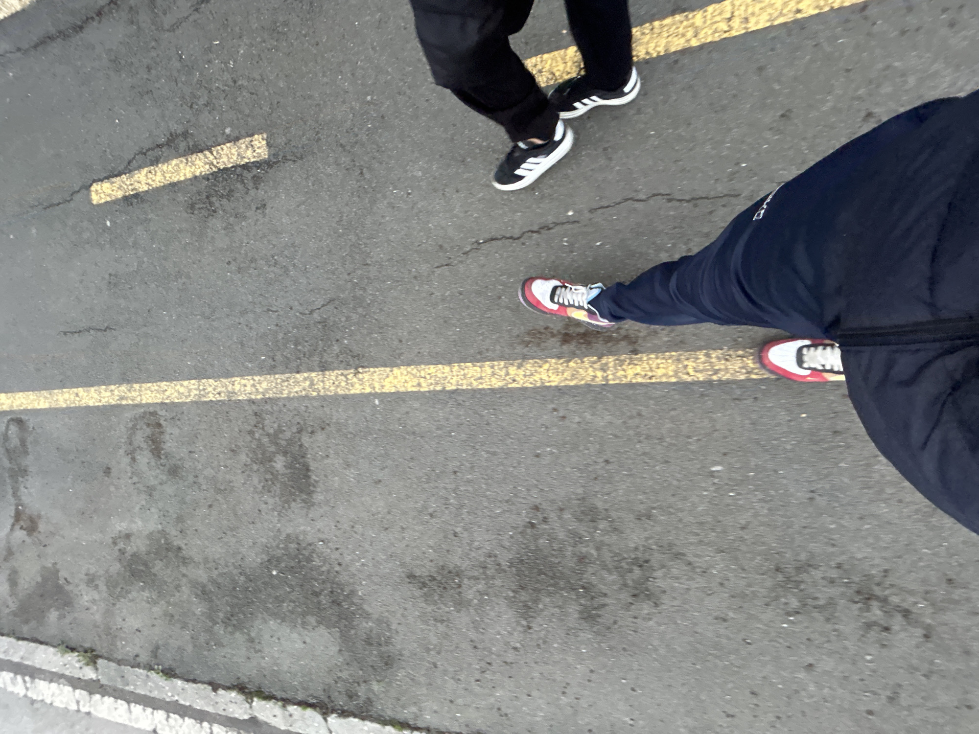 The image shows a section of a street in an urban setting. The surface is asphalt, marked with yellow lines. Two individuals are visible from the waist down. One person is wearing black pants and black and white shoes. Another person is wearing navy sweatpants and multi-colored sneakers. The location cannot be identified as Burgas, Bulgaria from the visible elements of the image.
