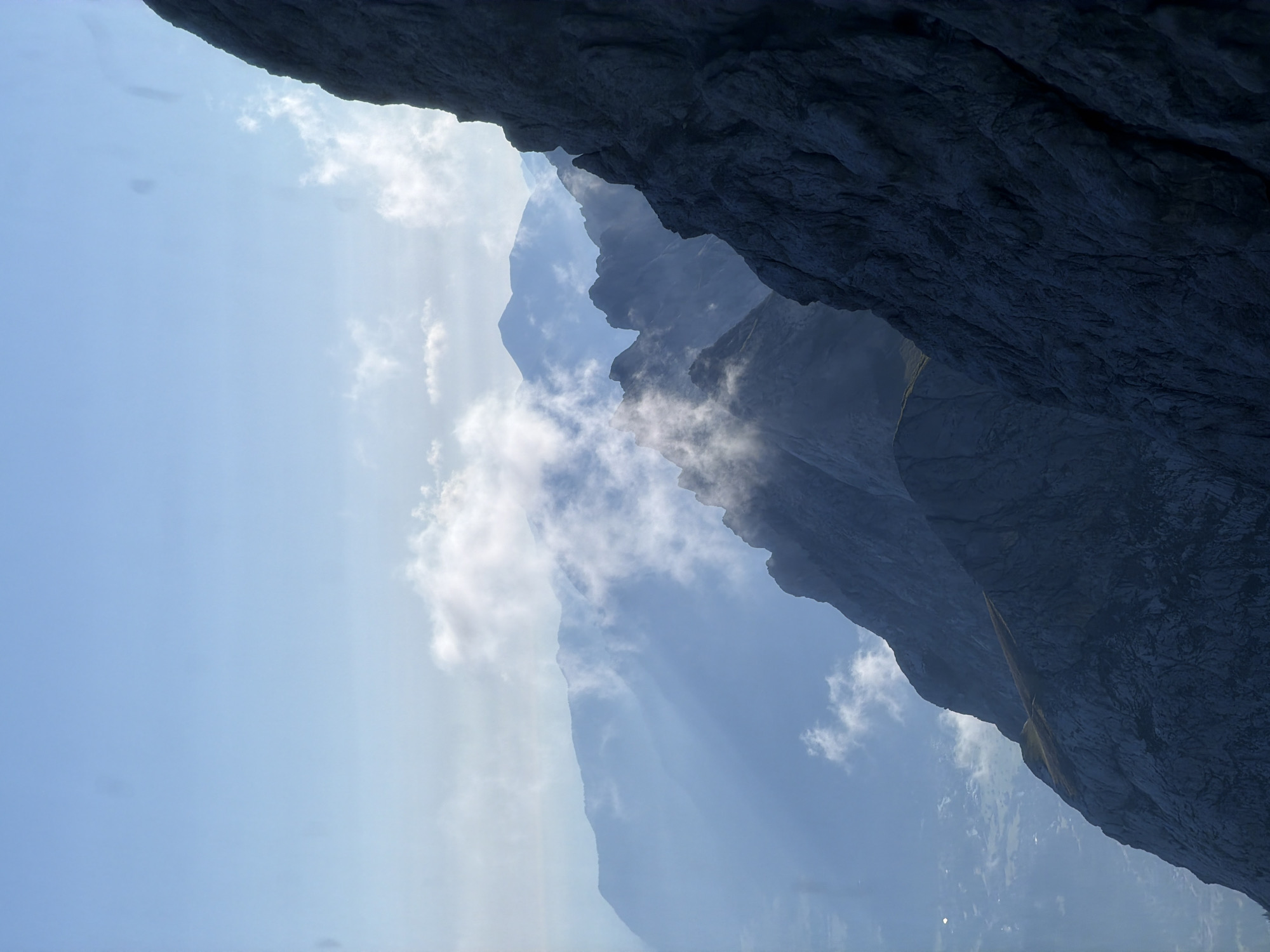 The image shows a dramatic mountain landscape in Grainau, Germany. The foreground is dominated by a steep, dark, rocky mountain face on the right side. In the middle ground, layers of jagged, blue-hued mountains recede into the distance, with wisps of white clouds and mist clinging to their peaks and slopes. Sunlight is casting shadows and highlights across the mountain ridges, creating a sense of depth and texture. The sky above is a clear, pale blue, with a few scattered, soft white clouds. The scene evokes a feeling of rugged natural beauty and remote wilderness. There are no people or man-made structures visible in the image.