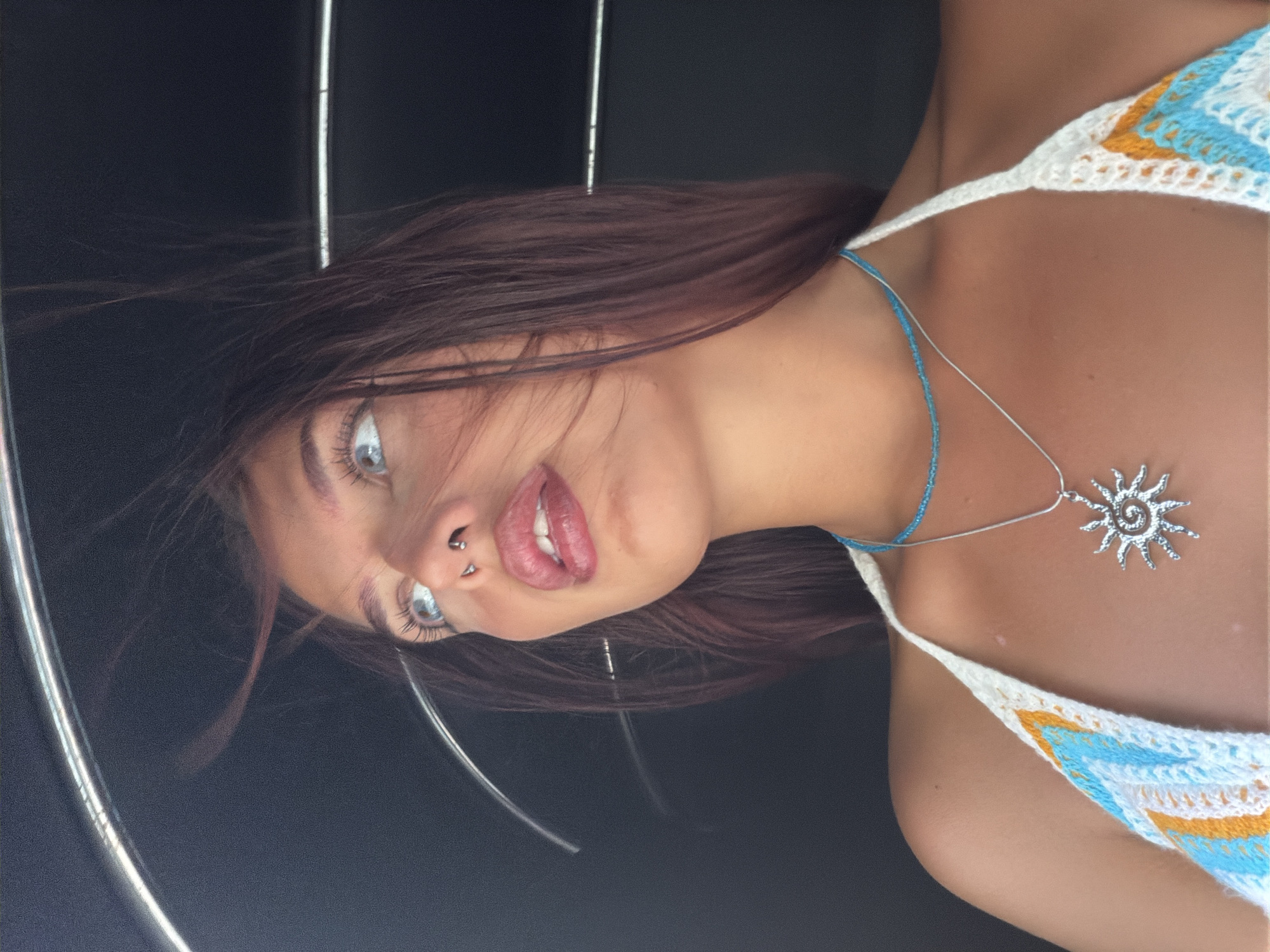 A young woman with striking blue eyes and a nose ring looks upwards and to her left. Her dark, reddish-brown hair is windswept. She is wearing a crocheted halter-neck top with a white, blue, and orange pattern, and a silver sun pendant necklace over a beaded blue necklace. The background is dark and appears to be the interior of a vehicle or structure with curved, metallic elements. The image was taken in Pottuvil, Sri Lanka.