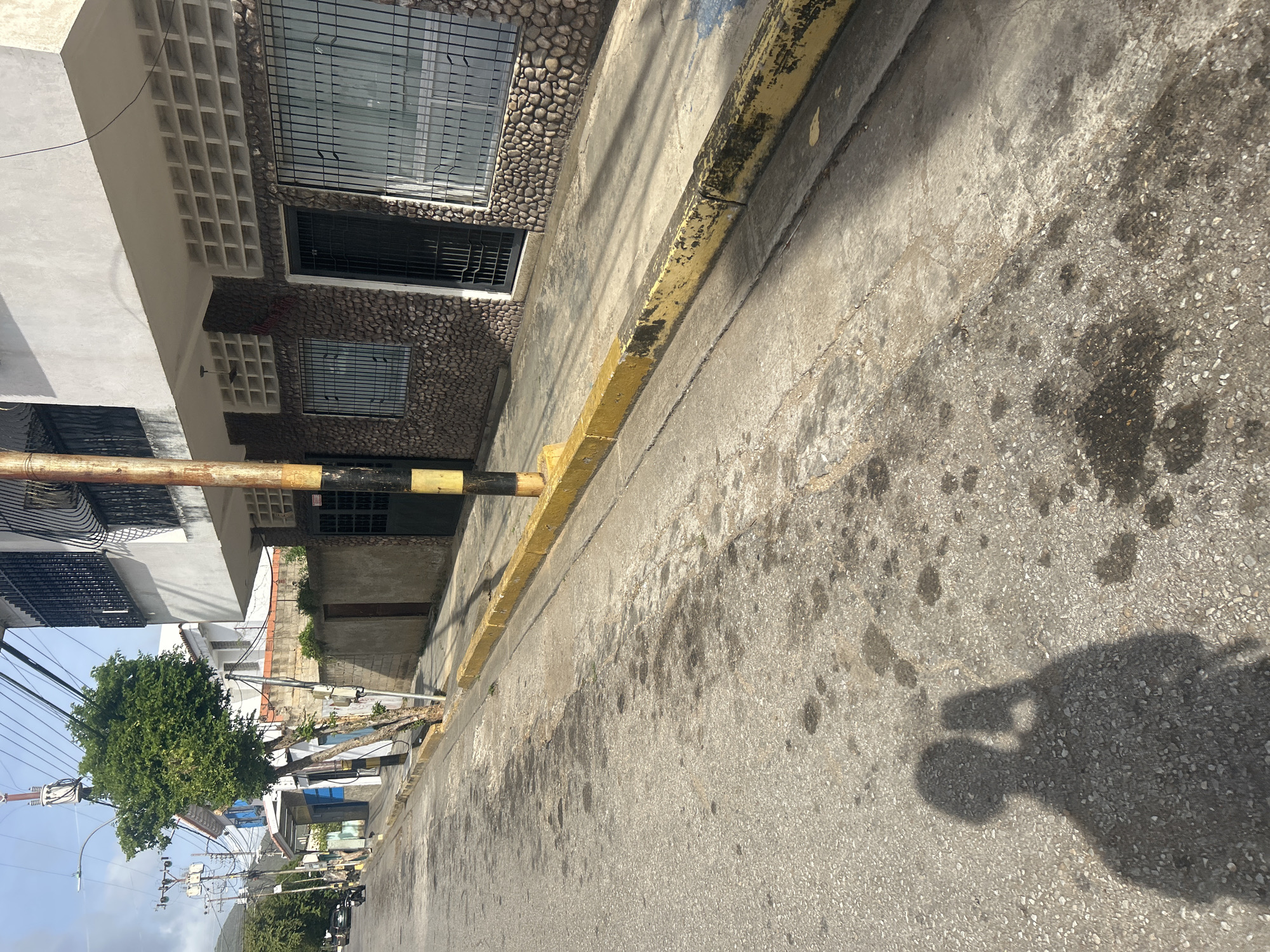 The image depicts a street scene in Santa Ana, Venezuela. A shadow of a person holding a phone is cast on the asphalt road in the foreground, suggesting a selfie was taken or a photo was being captured. The street is paved and shows signs of wear, with some dark patches that could be oil stains. A yellow-painted concrete curb lines the street, which is somewhat chipped and weathered.
To the right, a two-story building is visible. The ground floor is clad in small, rounded stones, and features black metal bars on windows and a black metal gate. Above the stone facade is a white section with white square ventilation blocks. The upper story is painted white. A black and yellow striped pole stands in front of the building.
To the left of the street, a sidewalk is present, with a small tree growing near the curb. Further down the street, more buildings and utility poles with many wires are visible. A motorcycle is parked on the left side of the street. The sky is partly cloudy. The overall impression is a moderately urban residential or commercial area with older infrastructure.