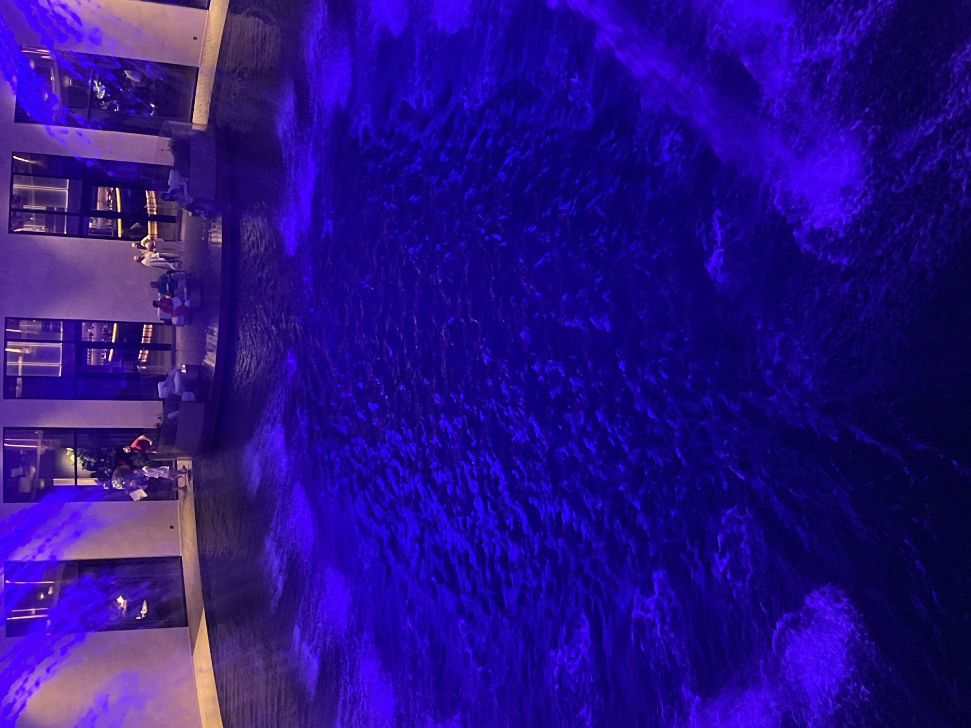 The image depicts an indoor swimming pool area, illuminated by vibrant blue lighting. In the background, a group of people are visible standing and sitting in a seating area with modern furniture, adjacent to a building with large windows. The water in the pool exhibits a textured surface with ripples and some areas of foam or splashes, likely from waterfalls or water features. The overall ambiance suggests a sophisticated establishment, possibly a hotel or a spa, located in the city of Krasnodar, Russia.