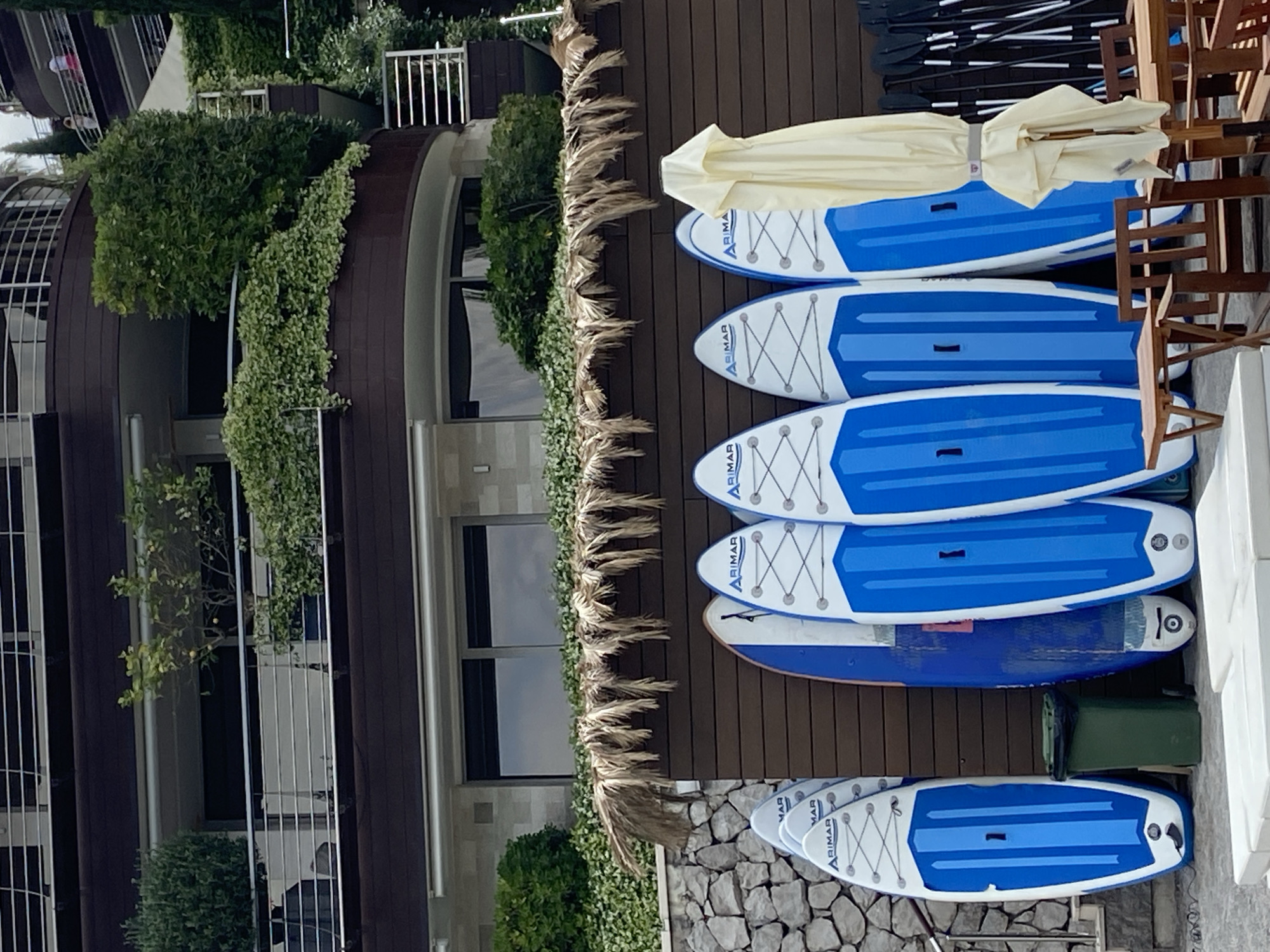The image shows a collection of paddleboards stored outside a building, likely near a waterfront area. The paddleboards are blue and white, stacked neatly on a wooden surface. Above the paddleboards, there is an umbrella and some wooden chairs. The building has a modern architectural style, with multiple floors and balconies covered in green plants.
Based on the scene details, especially the architecture, the location is likely Budva, Montenegro. Budva is a coastal city known for its modern buildings, lush greenery, and water activities. The paddleboards suggest a beach or water sports setting that is in line with this coastal destination.