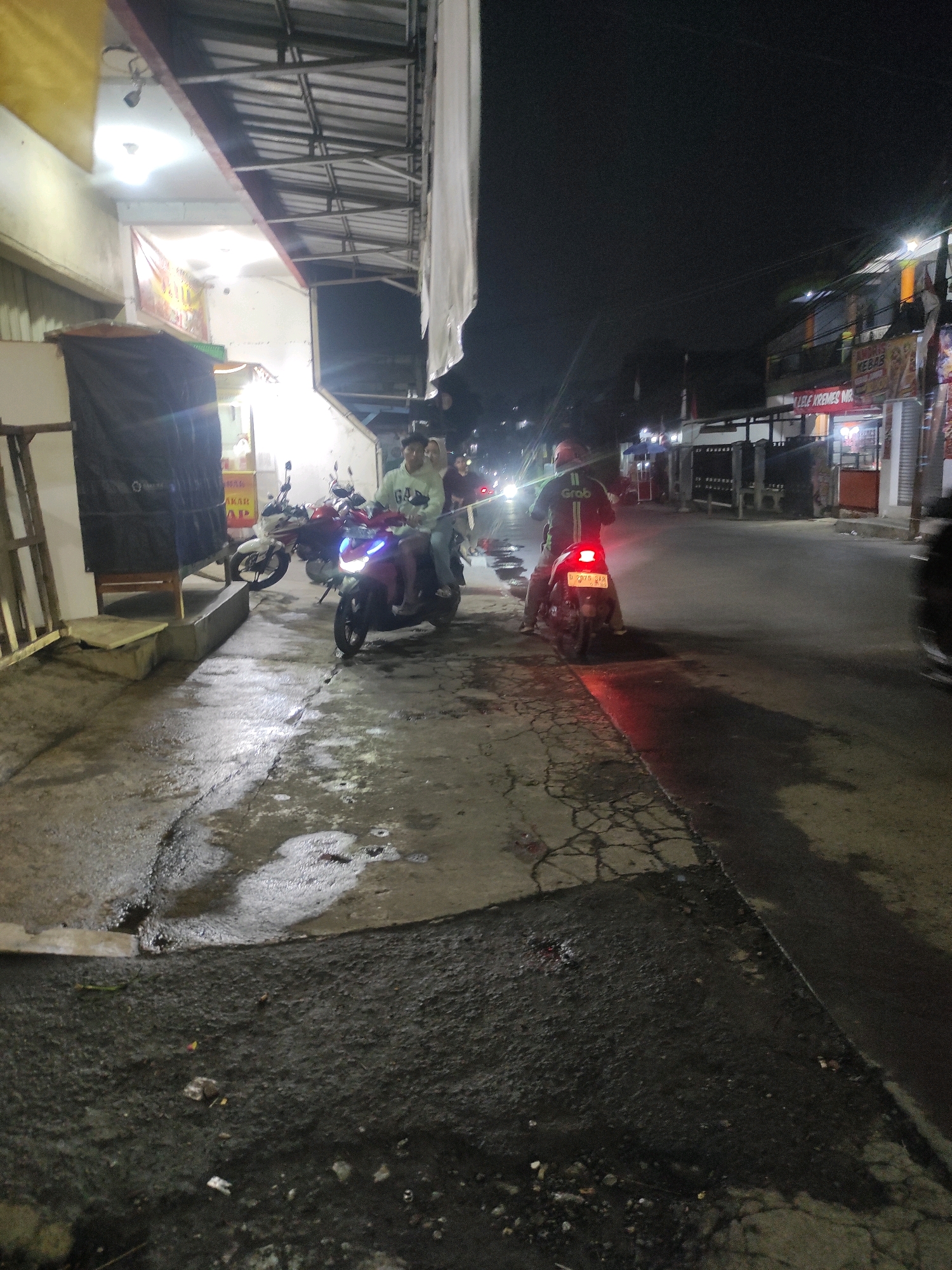 The image depicts a street scene at night in Padalarang, Indonesia, characterized by a wet pavement and dimly lit buildings. In the foreground, two individuals are seated on a red scooter, facing away from the camera. The rider wears a dark jacket with a white "Grab" logo on the back, suggesting they are a ride-hailing service driver. Another scooter, white and red, is parked behind them. Further down the street, another individual on a scooter, also facing away, is illuminated by the scooter's taillight, which casts a red glow on the wet pavement. The surrounding buildings appear to be shops or food stalls, with some illuminated signs visible. The overall atmosphere is that of a typical Indonesian street at night, with evidence of recent rain contributing to the wet and reflective surfaces.