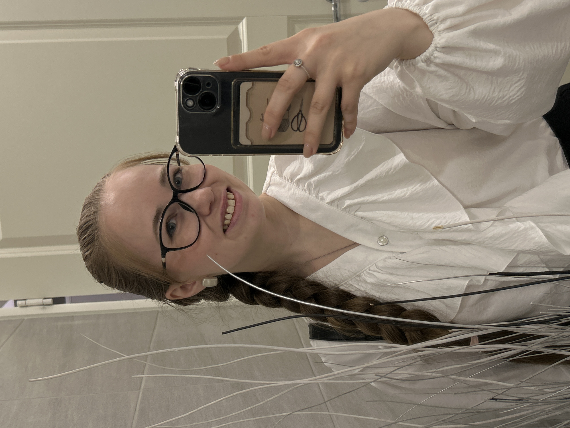 A young woman with light skin and reddish-blonde hair braided down her back is taking a selfie in a bathroom. She wears black-rimmed glasses and a white, button-down blouse with puffy sleeves. Her phone is held up in her right hand, revealing a clear case, a black phone body, and a card with a scissor design on the back. A ring is visible on her right ring finger. The background includes a white door, a tiled wall, and what appears to be decorative twigs. The location is unlikely to be in Saint Petersburg, Russia, based on the typical characteristics of bathrooms in that city.