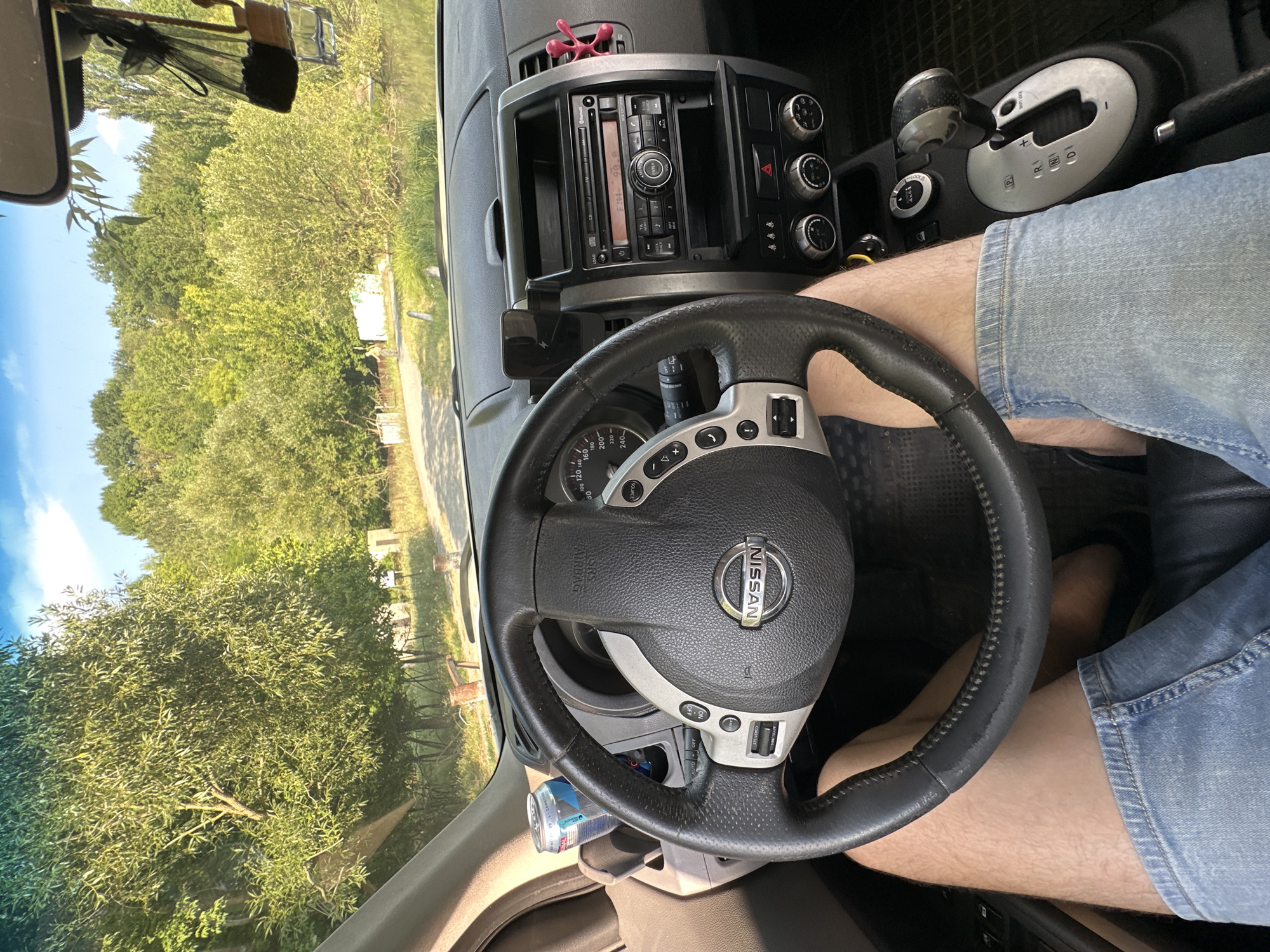 The image is a first-person perspective from inside a car, likely a Nissan X-Trail, parked on a dirt road in Makiv, Ukraine. The driver, whose legs and lower torso are visible, is wearing blue denim shorts. The car's interior is partially visible, including the steering wheel, dashboard with a radio and climate controls, and a gear shifter. Outside the car, a lush green landscape with trees and foliage is seen under a bright, partly cloudy sky. The road appears to lead towards a body of water and a wooded area. The setting suggests a rural or semi-rural environment.