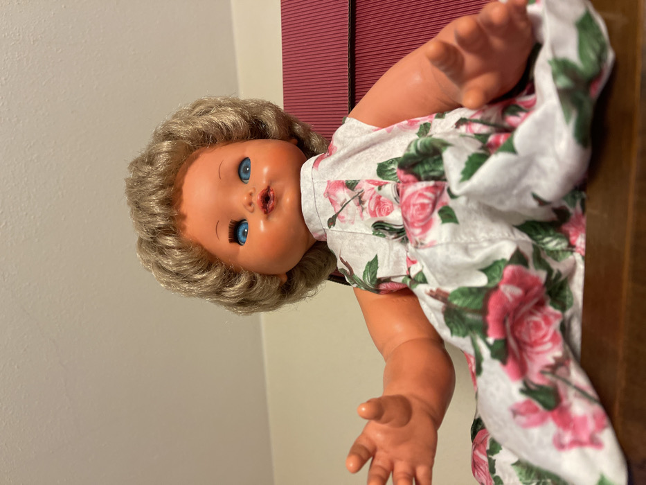 A doll is depicted in a close-up, angled shot, lying on its back with its head positioned towards the left of the frame. The doll has fair skin, bright blue eyes, and light brown, curly hair. Its mouth is slightly open, giving the impression of speaking or cooing. It is dressed in a white fabric garment, possibly a dress or romper, patterned with vibrant pink roses and green leaves.
The background to the left of the doll is a plain, light-colored wall, likely off-white or beige. To the right, behind the doll's body, is a darker, textured surface, appearing to be a piece of furniture or a blind with horizontal slats, in shades of reddish-brown or dark wood. The doll's arms and legs are visible, resting casually. The setting is indoors, with no specific activity occurring as the doll is static. There is no visible text in the image. The provided location context for the image is Torre di Santa Maria, Italy.
