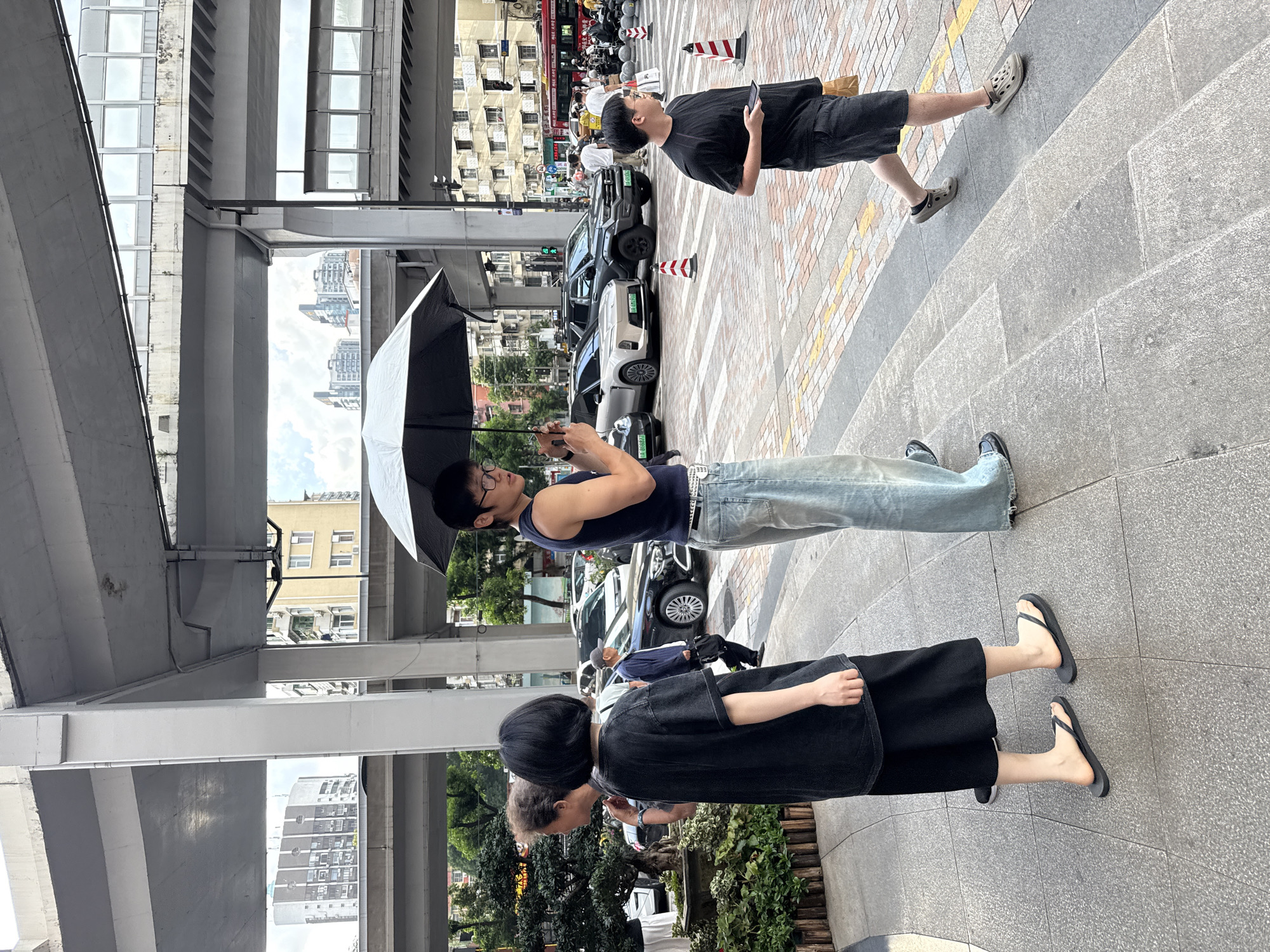 In Haizhu District, China, on a pedestrian walkway, four individuals are visible. In the foreground, a person with short black hair, wearing a black t-shirt and black skirt, is facing away from the camera, standing on flip-flops. To their left, another person's shoulder and arm are visible. A man holding an open black umbrella is in the center, wearing a blue tank top, light blue jeans, and glasses. To the right, a young person in a black t-shirt and dark shorts is walking away from the camera, holding a smartphone and a tan bag. The background features a busy street with several cars, including a silver sedan and a dark SUV, and buildings with multiple stories under an elevated road structure. Traffic cones and pedestrian crossing markings are present on the street. The overall lighting suggests it is daytime, possibly overcast.