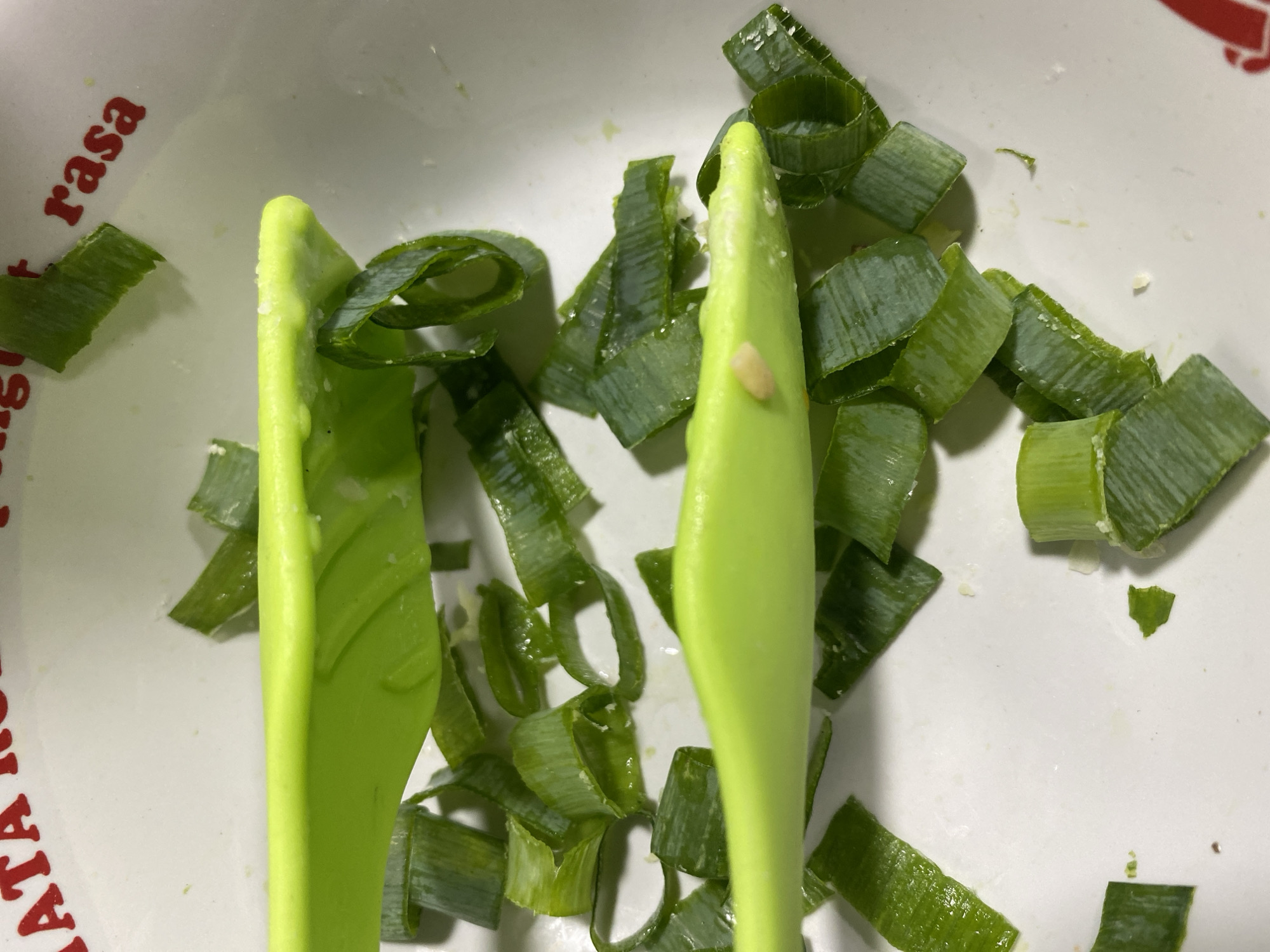 A close-up shot shows chopped green onions in a white bowl. The bowl has red text on its rim, which is partially visible. Two bright green spatulas are positioned across the bowl, with one partially covering the chopped onions and the other resting on top of them. The onions appear to be fresh and have been cut into various lengths. The lighting in the image is overhead, creating some reflections on the shiny surfaces of the onions and spatulas. The overall scene suggests food preparation. The text on the bowl, "RASA," implies the location is Indonesia, potentially within the city of Kasihan, known for its culinary traditions.