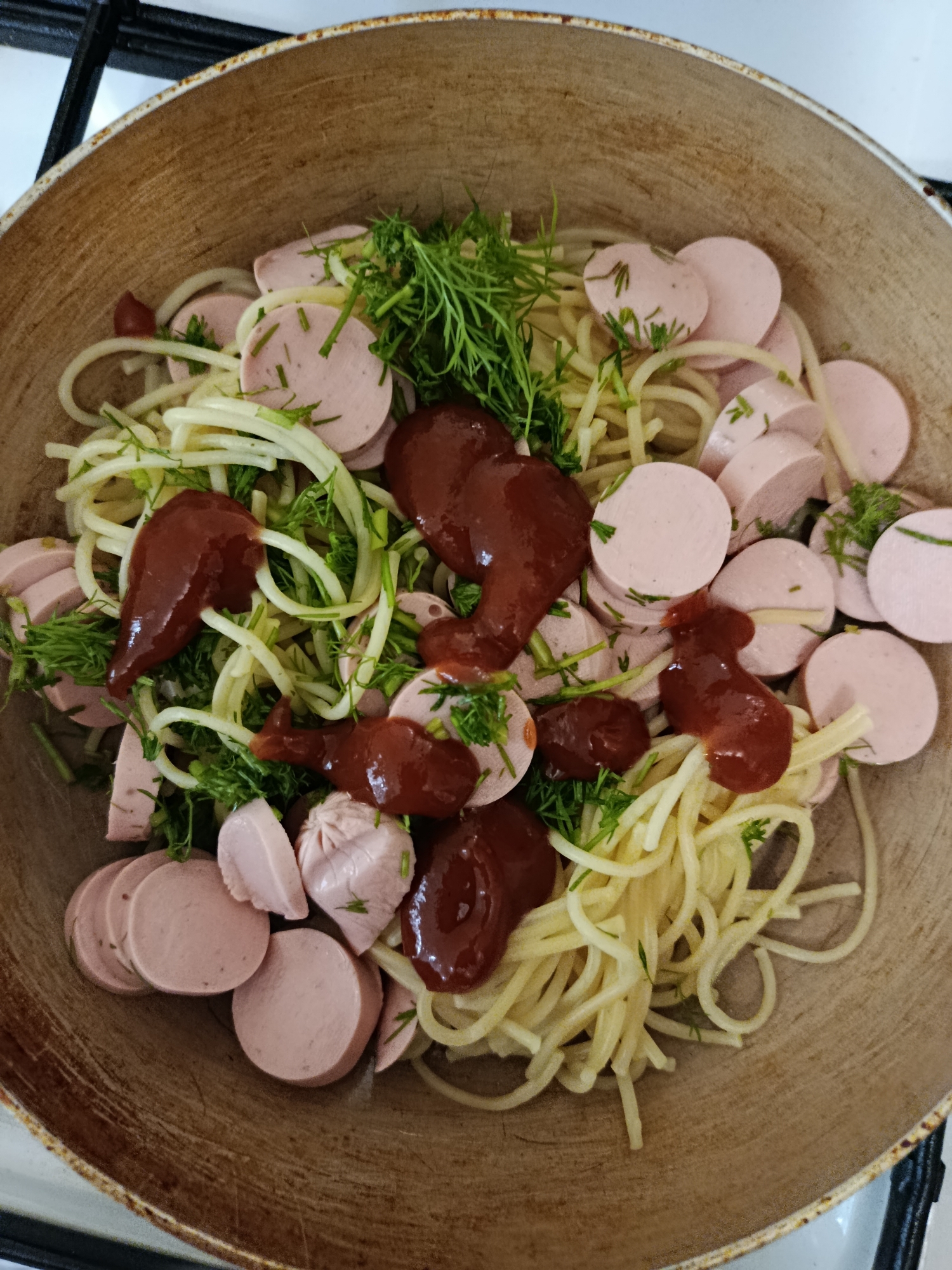 This is a close-up, overhead shot of a meal being prepared in a large, rustic ceramic pot or pan. The dish consists of spaghetti, sliced sausages, fresh dill, and dollops of dark red ketchup or tomato sauce. The spaghetti is a pale yellow and appears to be cooked al dente. The sausages are pinkish-white with visible red specks and are cut into thick, round slices, with a few pieces showing their pleated casing. The dill is bright green and finely chopped, scattered throughout the dish, adding a fresh aroma and visual contrast. The ketchup is distributed in several prominent dollops, some of which are slightly dripped and spread, suggesting it was recently added.
The setting appears to be a kitchen, indicated by the metallic stovetop visible in the background, which is slightly out of focus. The lighting suggests it's daytime, possibly indoors with artificial light supplementing natural light. There is no visible text in the image. The overall impression is of a simple, home-cooked meal being assembled or finished. The rustic pot and the casual presentation of the food suggest a comfortable, everyday setting. The meal looks hearty and potentially satisfying. The specific mention of Saratov, Russia, as the location context, suggests a domestic environment within that region.