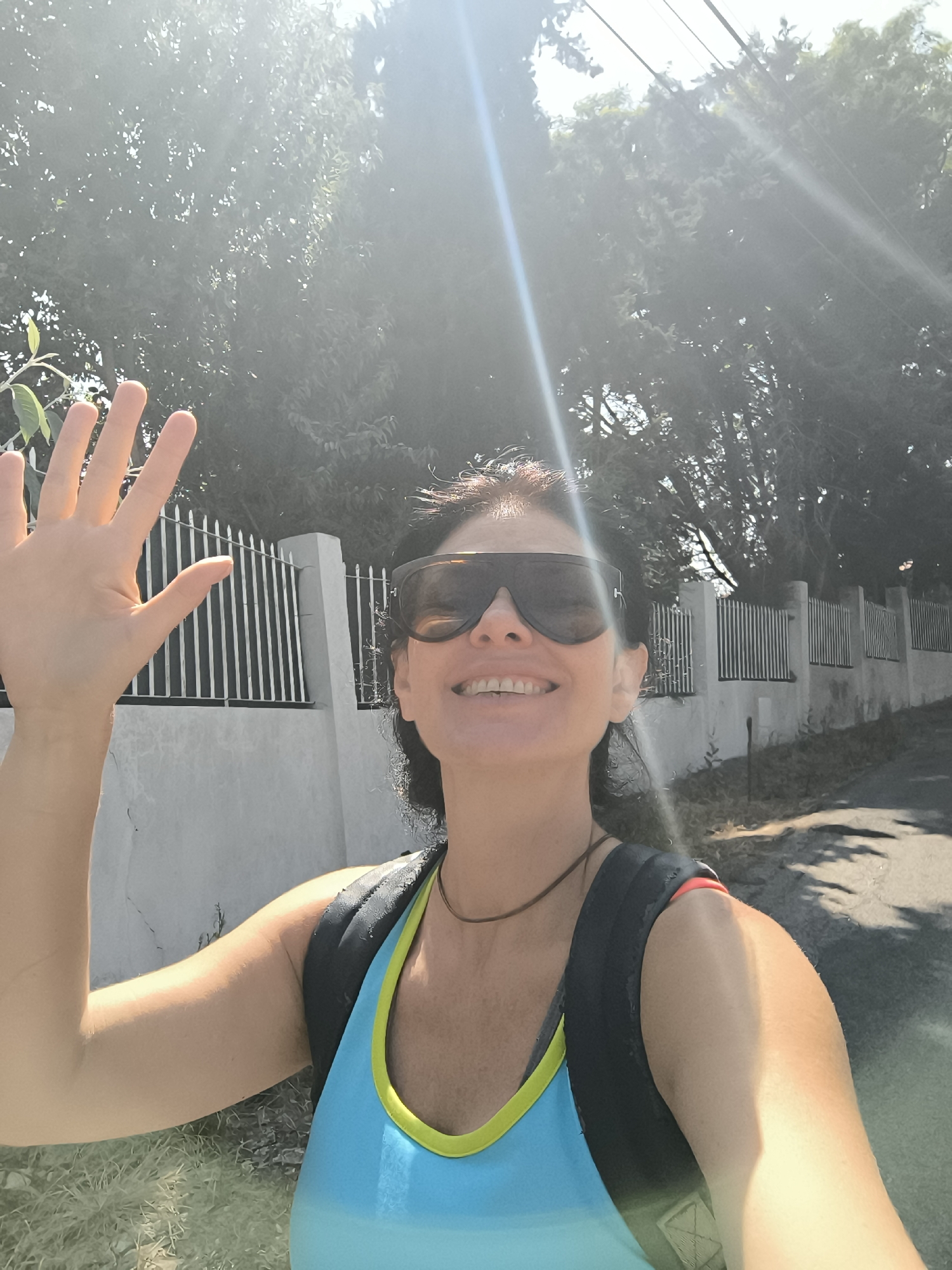 The image shows a woman with dark hair, wearing large sunglasses and a blue and yellow tank top. She is smiling and waving with her left hand, which is raised. She is also wearing a black backpack. The woman is standing on a paved road, with a white wall and a black metal fence behind her. Trees are visible in the background, and the sun is shining brightly, creating lens flares. The scene appears to be outdoors, possibly in a residential area. The location is identified as being in Palmela, Portugal.