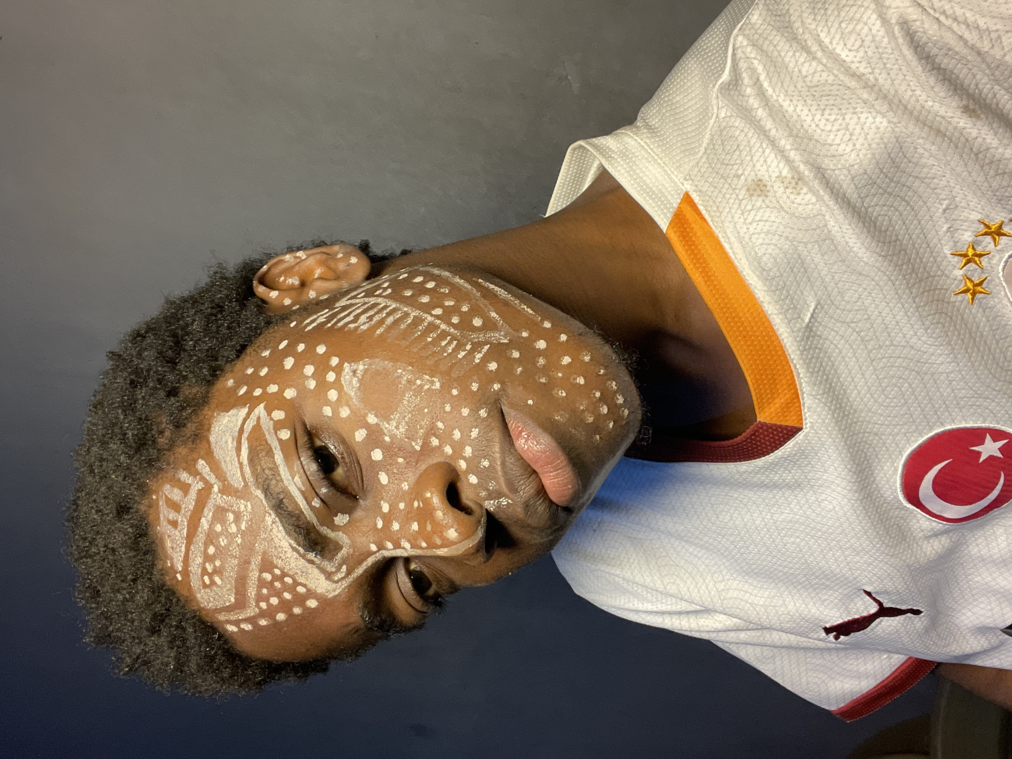 A person with dark skin and black curly hair is shown. The person's face is painted with white dots and lines in a tribal or decorative style. The person is wearing a white soccer jersey with orange and maroon accents. The jersey has a patch of the Turkish flag on the right chest and the Puma logo on the bottom. The image is a close-up shot, and the background is dark. The location of the image is not clearly identifiable as Nkpor, Nigeria.