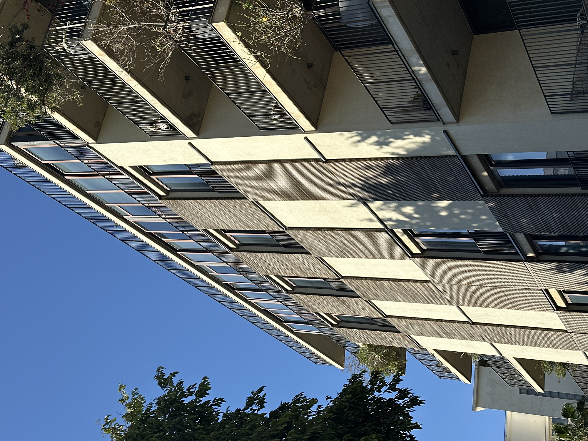 The image presents a striking view of a modern, multi-story building under a clear, bright blue sky. The architecture is characterized by a distinctive facade composed of alternating sections of light-colored, possibly concrete or stucco, walls and horizontal slatted screens, which appear to be made of wood or a similar material. These slatted elements create a pronounced play of light and shadow, with sharp, well-defined shadows cast across the building's surfaces, indicating intense sunlight.
The building features multiple balconies or recessed areas, each equipped with sleek horizontal metal railings. Within some of these balconies, small planters hold sparse, dry-looking vegetation, suggesting a resilient flora. At the bottom of the frame, the vibrant green foliage of trees adds a touch of nature to the urban environment. The windows of the building are reflective, mirroring the expanse of the blue sky above.
The overall aesthetic is contemporary, with a focus on geometric patterns and clean lines. The bright sunlight and deep shadows suggest the photograph was taken on a sunny day, likely during the mid-morning or afternoon. There are no people visible, and no specific activities are occurring. No text is discernible in the image. This scene is situated in Tel-Aviv, Israel, reflecting the city's modern architectural landscape.