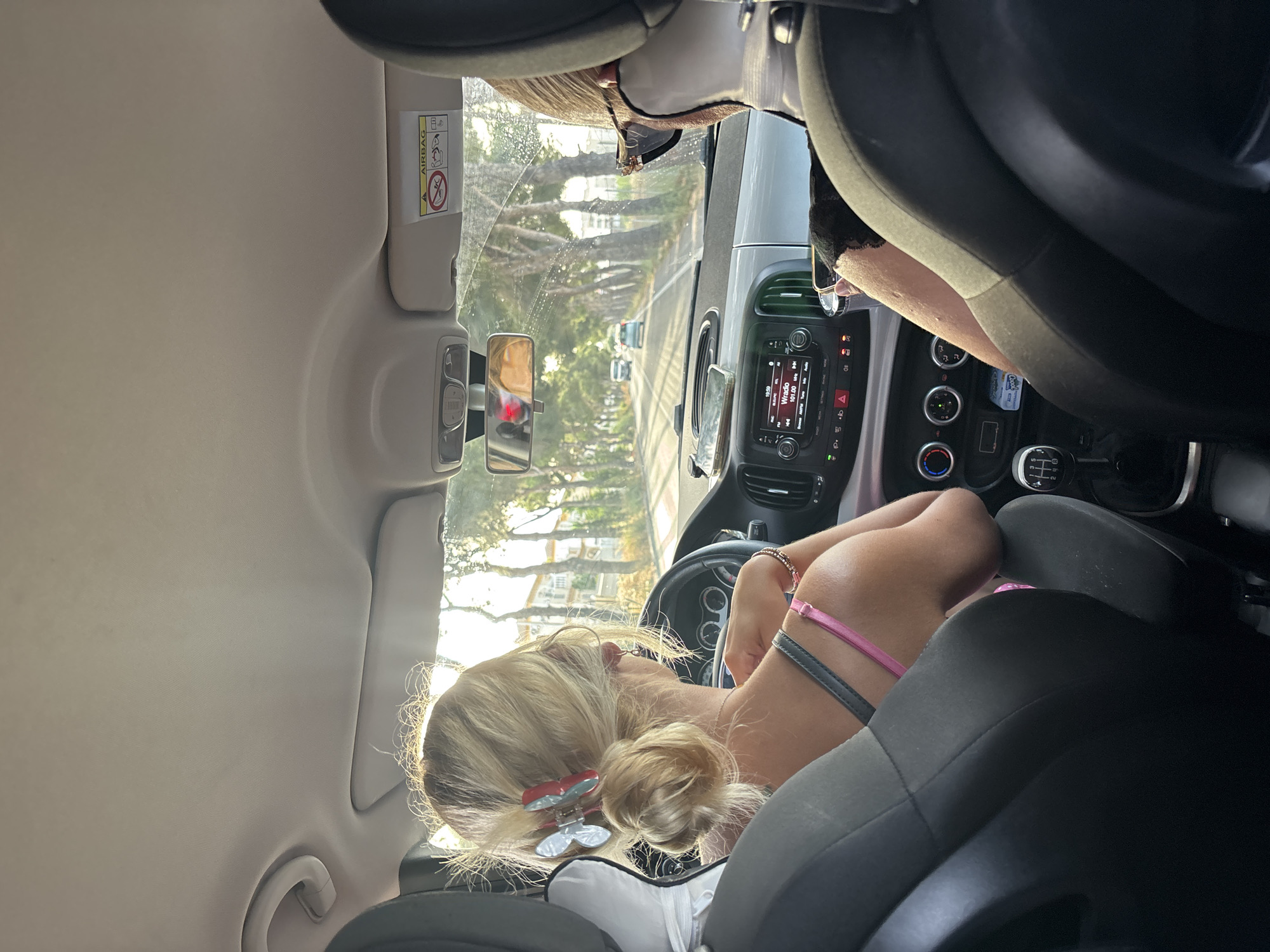 In the city of Orihuela, Spain, two individuals are captured from behind, seated inside a vehicle moving along a tree-lined road. The driver, a blonde woman with her hair styled in a bun and adorned with butterfly hair clips, wears a pink top. Her hands are on the steering wheel, suggesting she is actively driving. Beside her, a second passenger, whose face is obscured by large sunglasses and a face mask, holds a phone. The car's interior is visible, including the dashboard with its controls and a car radio displaying "WrRadio." Outside the car, the road curves gently, with the sun casting a warm glow through the foliage of the trees lining the route. Several other vehicles are visible in the distance. The overall scene depicts a casual journey through the Spanish landscape.