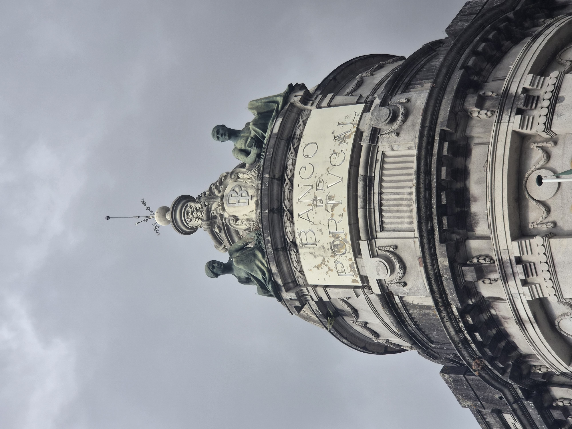 The image shows the facade of the Banco de Portugal building in Braga, Portugal, under a cloudy sky. Two statues of seated figures, one male and one female, flank a central emblem above the words "BANCO DE PORTUGAL" carved into the stone. The architecture is classical, with decorative elements like garlands and fluted columns visible. The stone appears weathered, with some areas showing peeling paint or grime. A flagpole with a white pole and a green flag is visible at the bottom center of the frame.