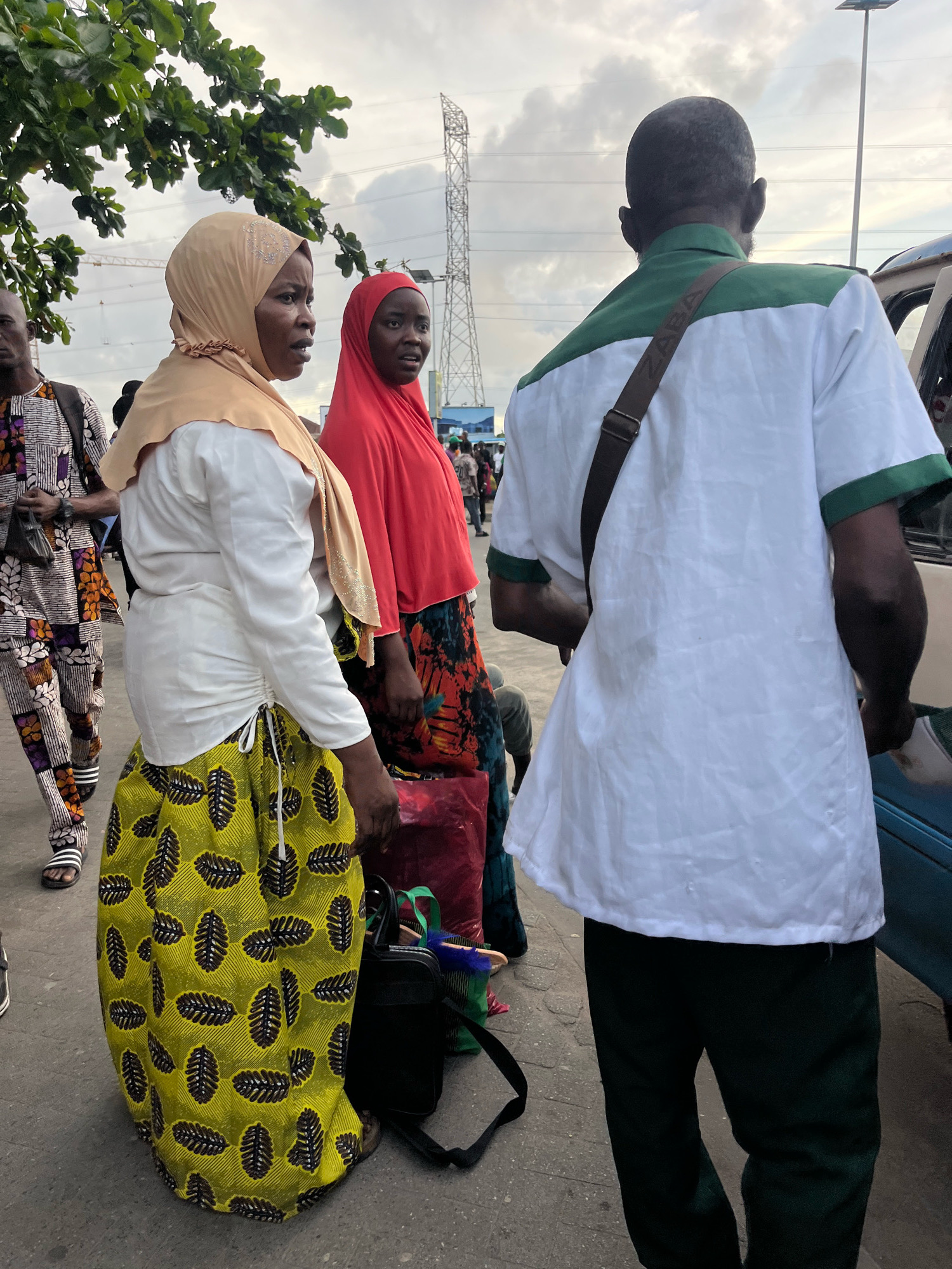 In Lekki Phase I, Nigeria, a group of people are gathered outdoors, possibly at a transportation hub, under an overcast sky. A man in the foreground, facing away from the camera, wears a white short-sleeved shirt with green trim on the collar and cuffs, and dark green trousers. He has a brown strap across his chest. To his left, two women are visible. One woman wears a light tan headscarf with embellishments around the edge and a white blouse, paired with a yellow and black patterned skirt. The other woman to her left wears a bright red headscarf and a patterned dress. The background shows trees, a tall tower, and what appears to be a blue vehicle. Other individuals and bags are partially visible, suggesting a busy environment.