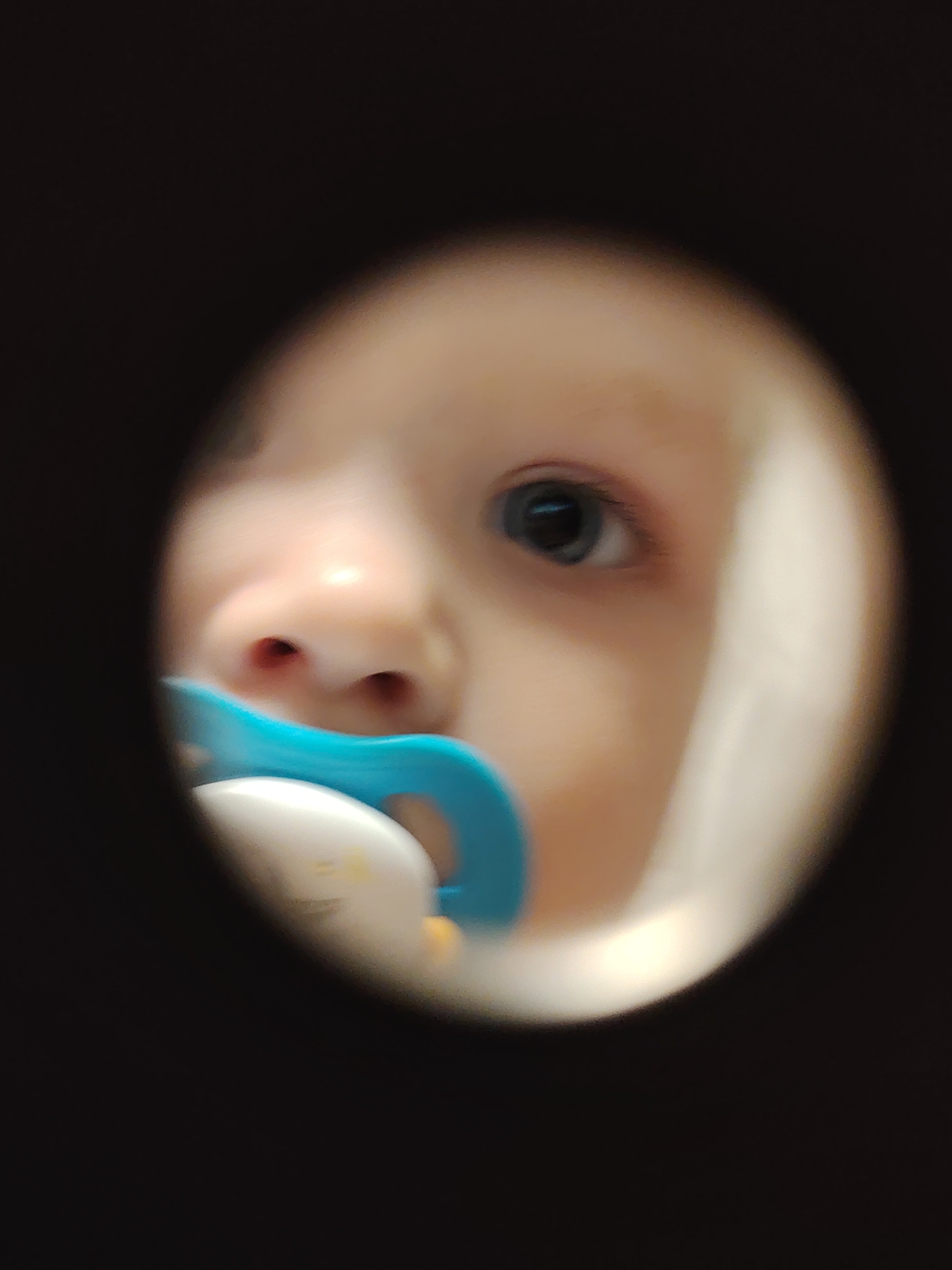 A close-up shot of a baby's face, looking through a circular opening. The baby has fair skin, blue eyes, and is sucking on a blue and white pacifier. The surrounding area is dark and out of focus, creating a peephole effect. There is no information available about this image being located in the city of Chaikovsky, Russia.