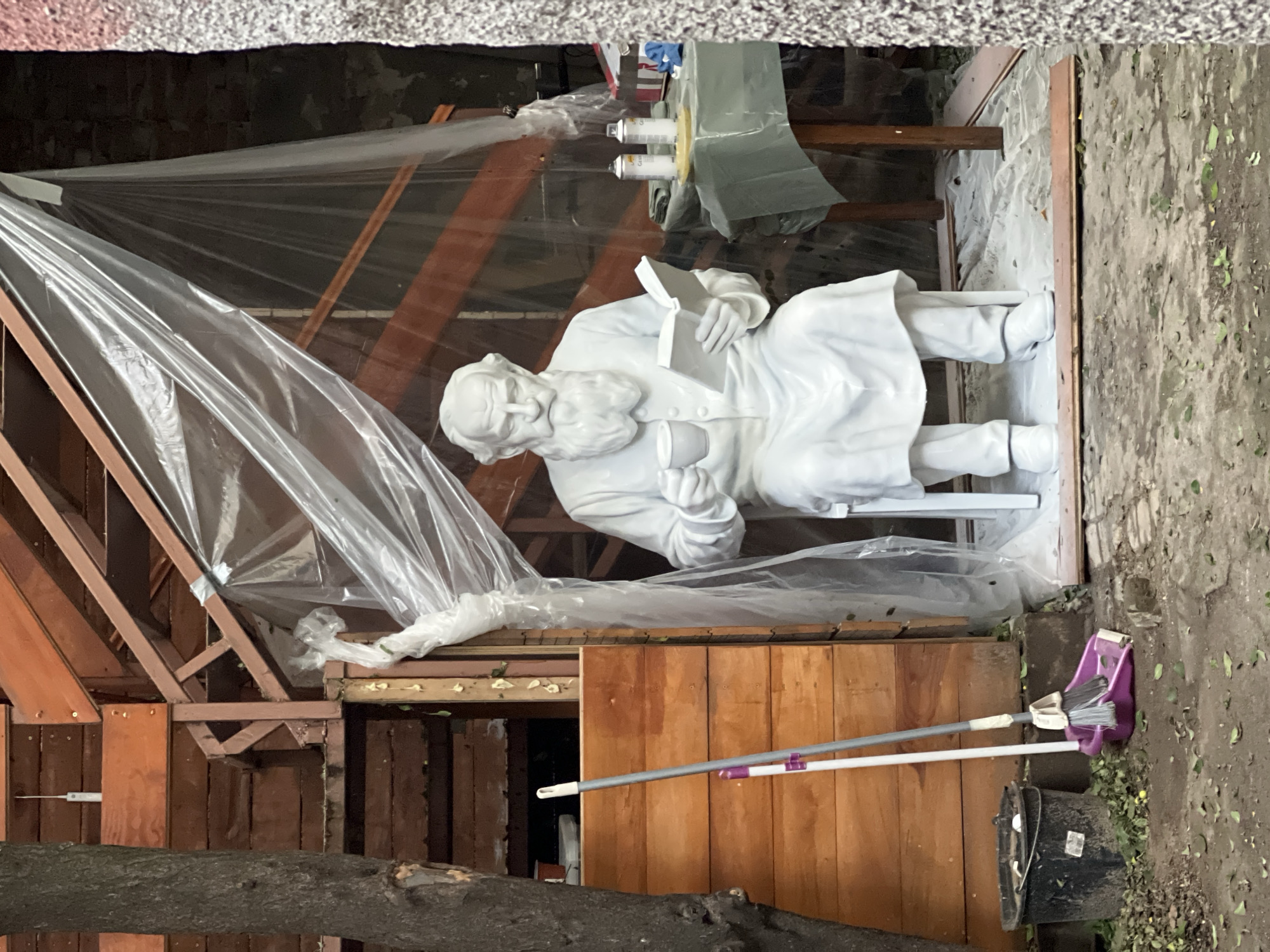 An outdoor scene in Plovdiv, Bulgaria, depicts a freshly painted white statue of a bearded man seated and reading a book. The statue is located inside of a partially enclosed wooden structure covered with a clear plastic sheet. Two cans of white spray paint sit on a small wooden table behind the statue. A broom and dustpan are placed against the side of the enclosure near a black bucket. The ground is dirt and scattered leaves.