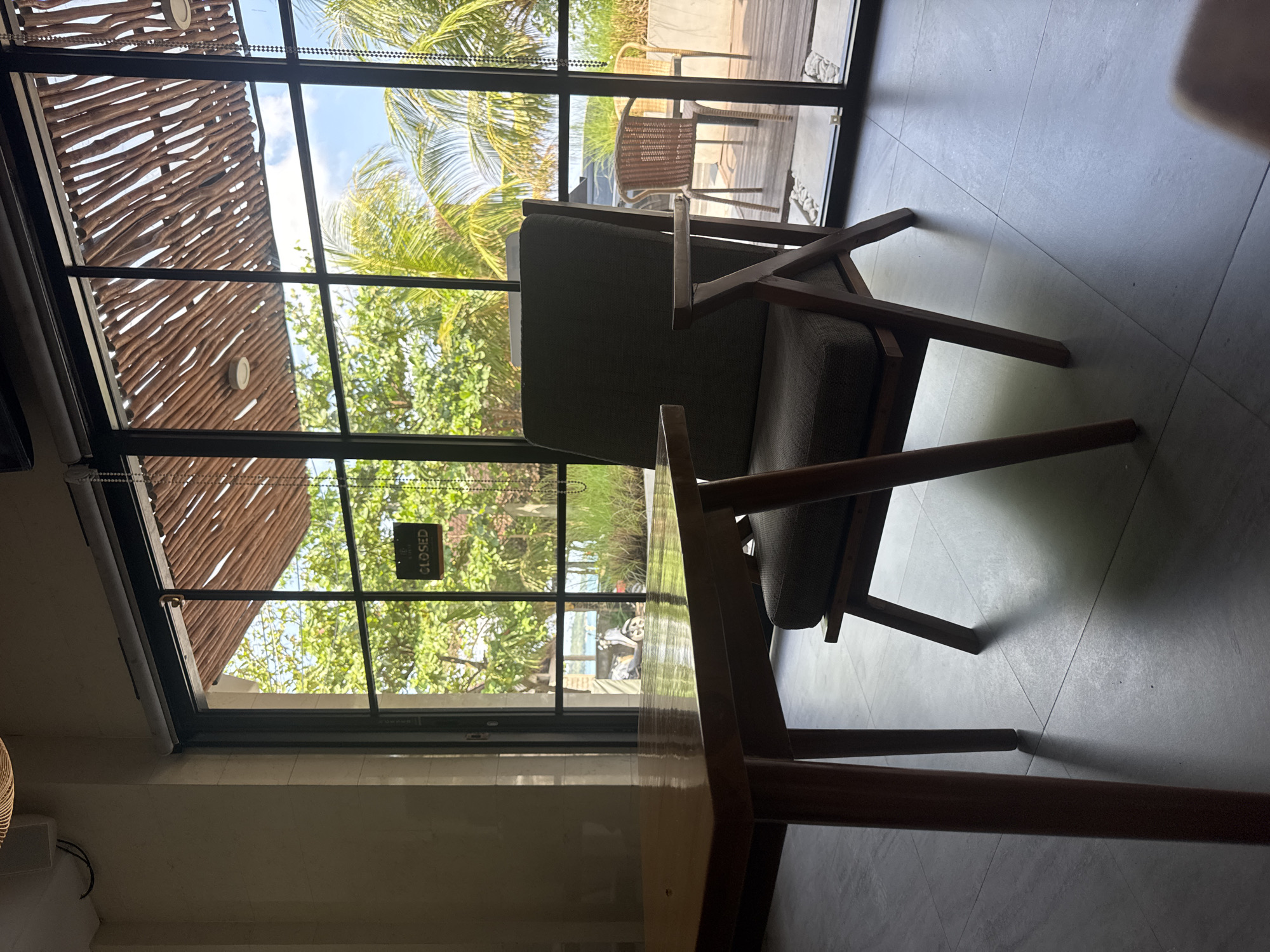 The image shows the interior of a cafe or restaurant, likely in Kuta, Indonesia. The primary subjects are two chairs. One is a modern design with dark gray cushions and a wooden frame, partially visible against a large window. Outside the window, lush green vegetation, possibly palm trees, is visible, suggesting a tropical environment typical of Bali. A glimpse of another chair outside on what appears to be a wooden deck can be seen through the window. The second chair is a wooden table, obstructing the camera. The interior has a modern aesthetic with tiled floors. A "CLOSED" sign hangs on the window, indicating the establishment is not currently open. The lighting suggests it is daytime, but the interior has a somewhat dim atmosphere.