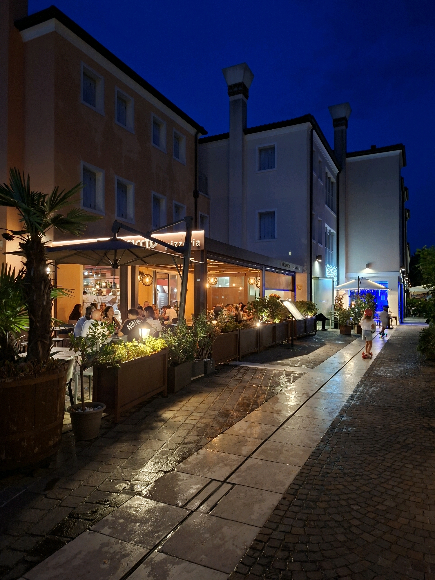 A street scene in Caorle, Italy, at dusk. A restaurant with outdoor seating is illuminated by warm lights, casting reflections on the wet cobblestone street. People are seated at tables, some enjoying meals, others in conversation. A palm tree stands prominently on the left side of the frame. Two children are visible in the distance, one on a scooter and another walking. The buildings flanking the street are two and three stories high, with windows and architectural details typical of Italian urban environments. The sky is a deep blue, indicating the late hour.