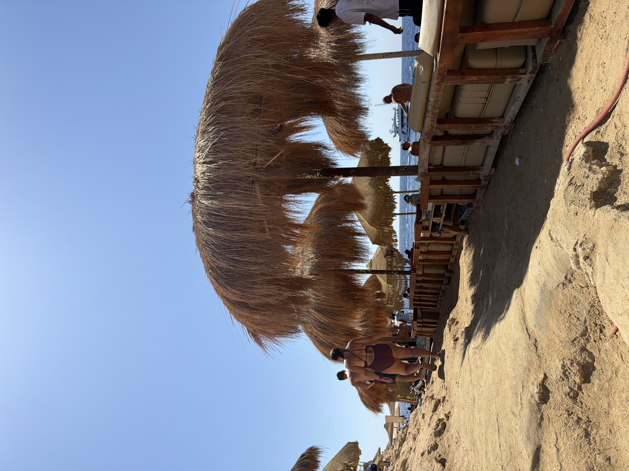 A sunny beach scene in Sharm El Sheikh, Egypt, features a sandy pathway leading towards the sea. Large thatched umbrellas provide shade over seating areas constructed from wooden frames and cushions. Several people are visible, some walking along the path, others relaxing under the umbrellas or in the water. A boat is present in the distance on the clear blue sea. The environment is characterized by sand, sun, and calm water.