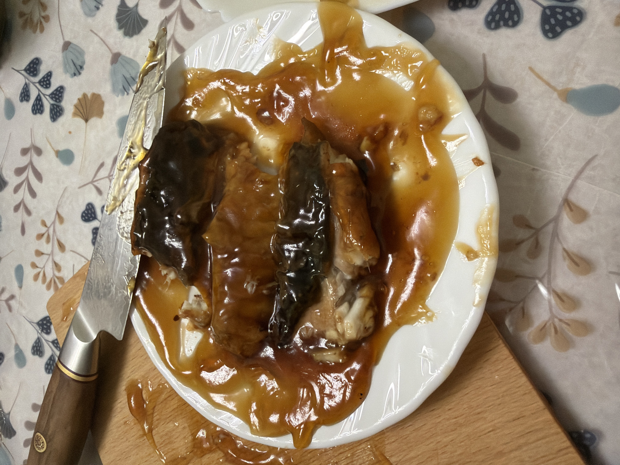 A close-up shot shows a white plate with pieces of unagi, covered in a glossy brown sauce, resting on a patterned tablecloth. A knife with a wooden handle rests on the plate, its blade coated in the same sauce. A portion of a wooden cutting board is visible to the left of the plate. The scene is illuminated by overhead lighting, casting shadows on the tablecloth. The background features a floral pattern in muted colors of blue, brown, and pink.
