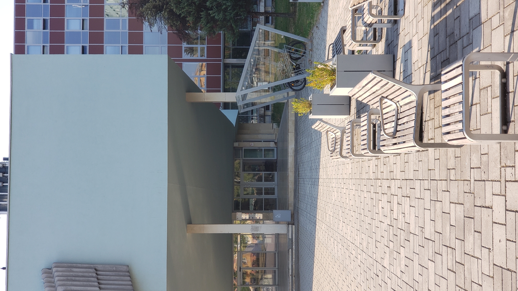 The image shows a modern building with a light blue facade and a red-brown section with many windows. In front of the building is a paved courtyard with a brick-like pattern. Several modern benches are arranged in a row on the paved area, with some of them casting shadows. There are also planter boxes with small green plants. To the right, there is a contemporary glass and metal structure that appears to be a bicycle rack, with bicycles inside. A mature tree is visible behind the bicycle rack. The entrance to the building features glass doors and is covered by an overhang supported by columns. The overall scene suggests a public space in an urban environment, possibly near a university or office building. Based on the architecture and the context of Prešov, Slovakia, this location is the Faculty of Arts of the University of Prešov, located at Ulica 17. novembra 1.