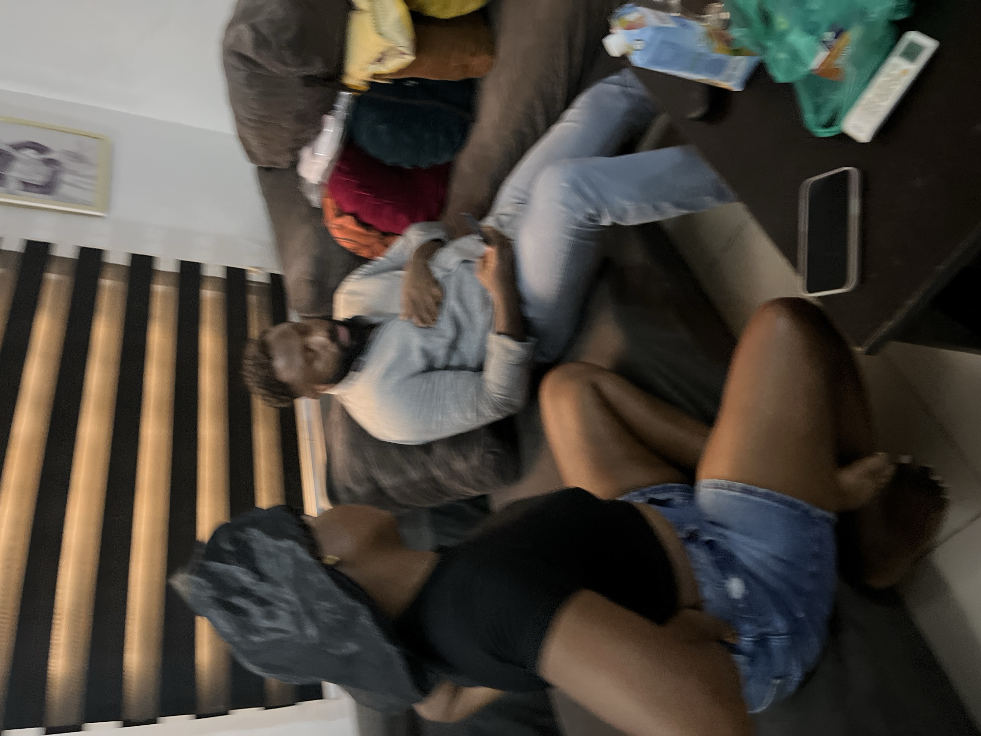 In a dimly lit room, possibly a living room in Abuja, Nigeria, a woman wearing a black t-shirt and denim shorts sits on a couch, her back to the camera. Her hair is covered with a black bonnet. A man, also on the couch, is wearing a light blue button-down shirt and light blue pants. He is looking down at his phone. Several pillows in various colors are scattered on the couch behind him. A black coffee table in the foreground has a smartphone, a remote control, and a carton of juice on it. The room is decorated with blinds on the window and a framed picture on the wall.