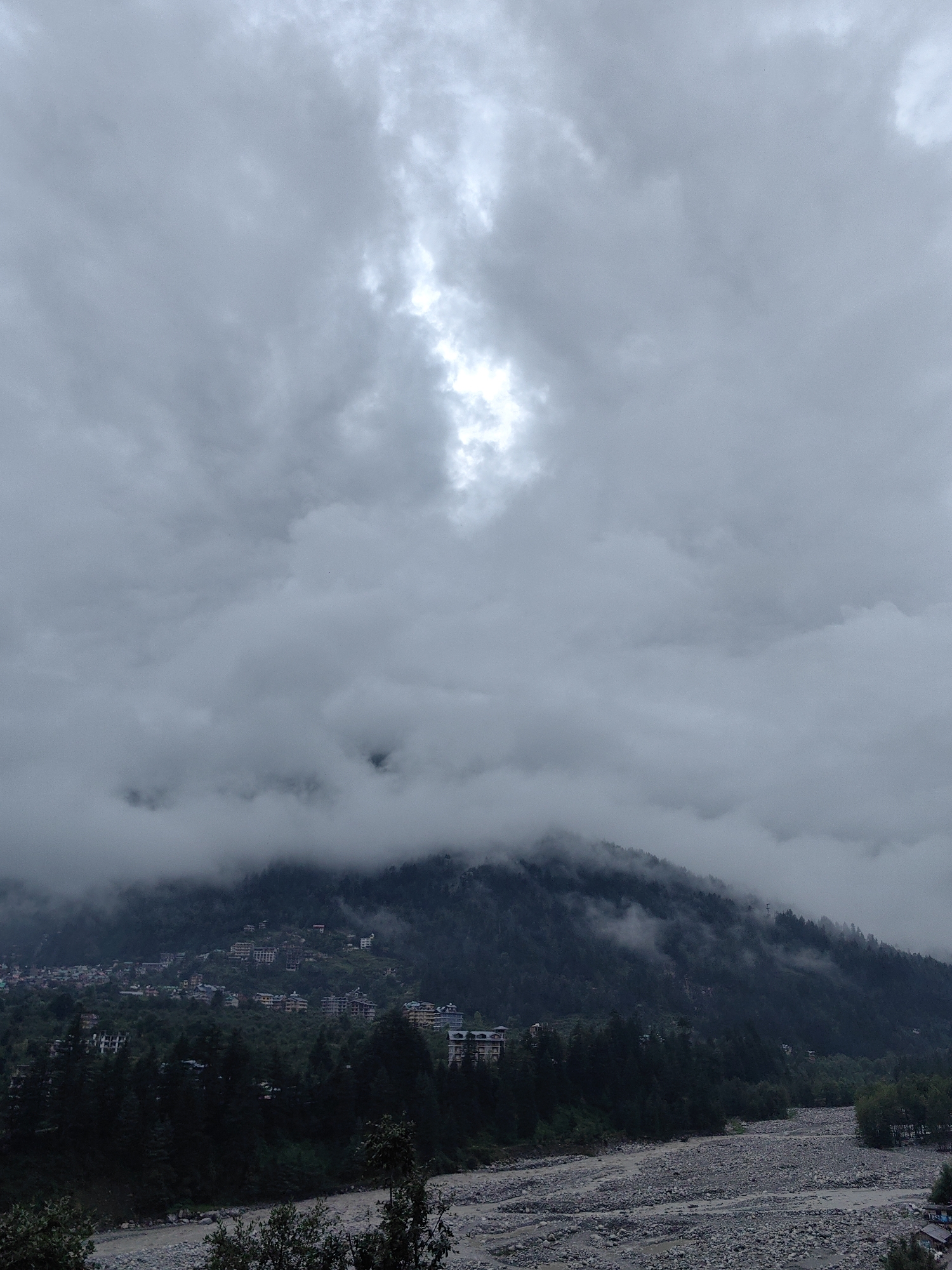 The image captures a cloudy, possibly overcast day in a mountainous region. The scene is dominated by a large, tree-covered mountain that slopes down towards a rocky riverbed. Numerous buildings, varying in size and architectural style, are scattered across the mountainside, suggesting a settlement or town. The buildings appear to be residential or commercial, with some resembling hotels or apartment complexes.
The sky is filled with thick, grey clouds, with a bright opening revealing sunlight filtering through in the upper central portion of the image. This break in the clouds creates a dramatic contrast against the otherwise muted tones of the landscape. The mountains are shrouded in mist and low-lying clouds, particularly around their peaks, adding to the atmospheric and perhaps somewhat mysterious quality of the scene.
In the foreground, a wide riverbed filled with stones and pebbles is visible. The water level appears low, with most of the riverbed exposed. Lush green trees, predominantly pine or similar coniferous species, form a dense forest along the lower slopes of the mountain and the banks of the river.
The overall mood of the image is somber and serene, possibly evoking a sense of peacefulness or even a hint of melancholy due to the cloudy weather. The location appears to be a developing area with a mix of natural beauty and human infrastructure. There is no visible text in the image.