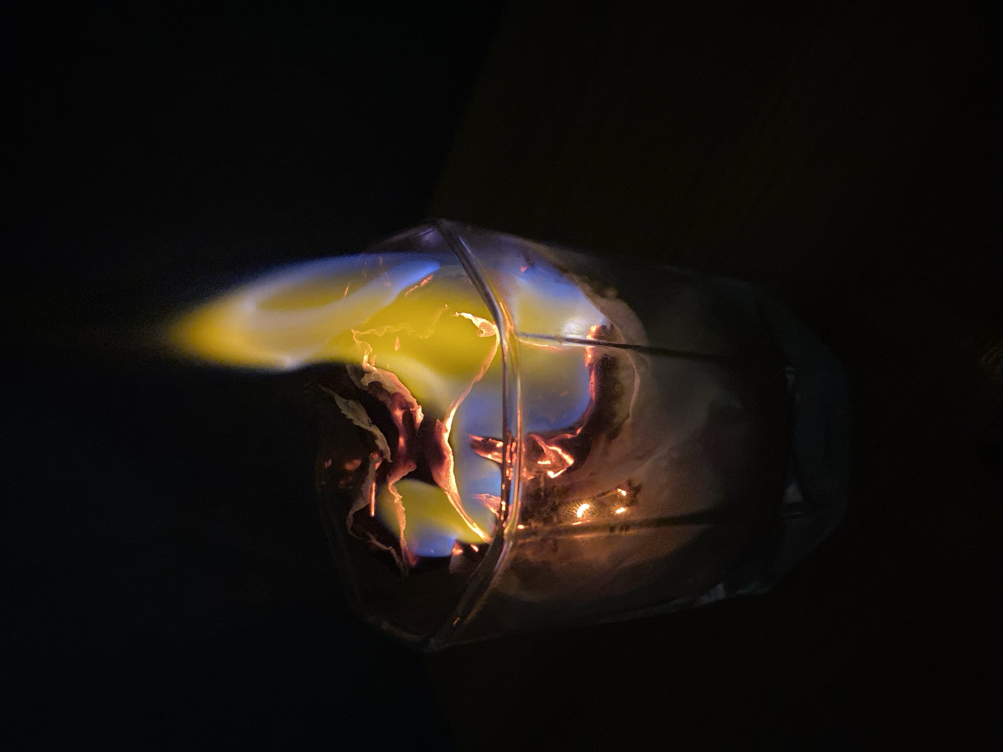 The image depicts a fire contained within a clear, geometric structure resembling a lantern or cage. The flames are predominantly yellow, with blue hues near the base, and extend outside of the structure. The background is completely dark. The city location is unknown.