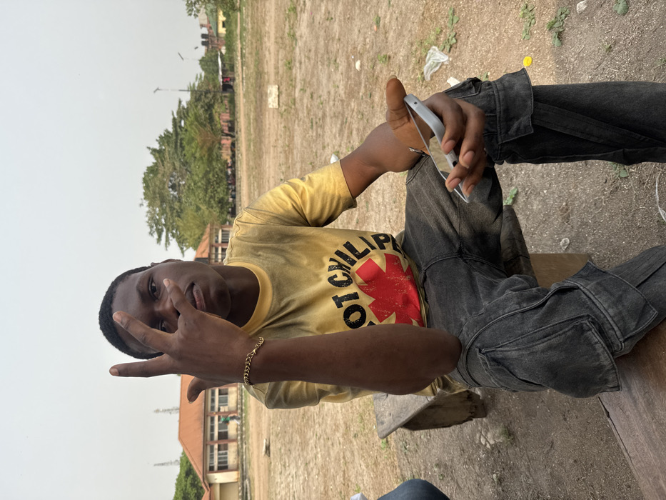 A young male subject with dark skin is seated on a weathered wooden bench in an open, undeveloped area within Ihiagwa, Nigeria. He wears a yellow, tie-dye short-sleeved t-shirt with a red asterisk-like logo and the partial text "HOT CHILI" printed on the front, dark grey cargo pants, and a gold-colored chain bracelet on his left wrist. His right hand is raised to chest height, with the index and middle fingers extended upward and the thumb, ring, and pinky fingers bent. His left hand holds a white smartphone horizontally. The subject is looking directly at the camera. The ground is dry, dusty earth with sparse green vegetation and scattered litter, including plastic bags and a yellow cap. In the background, several light-colored, low-rise buildings with reddish-brown tiled roofs are visible, consistent with institutional or academic structures, bordered by large green trees and utility poles. The sky is overcast. A segment of a person's leg, clad in blue jeans, is visible on the far left of the bench, indicating another individual is seated nearby.