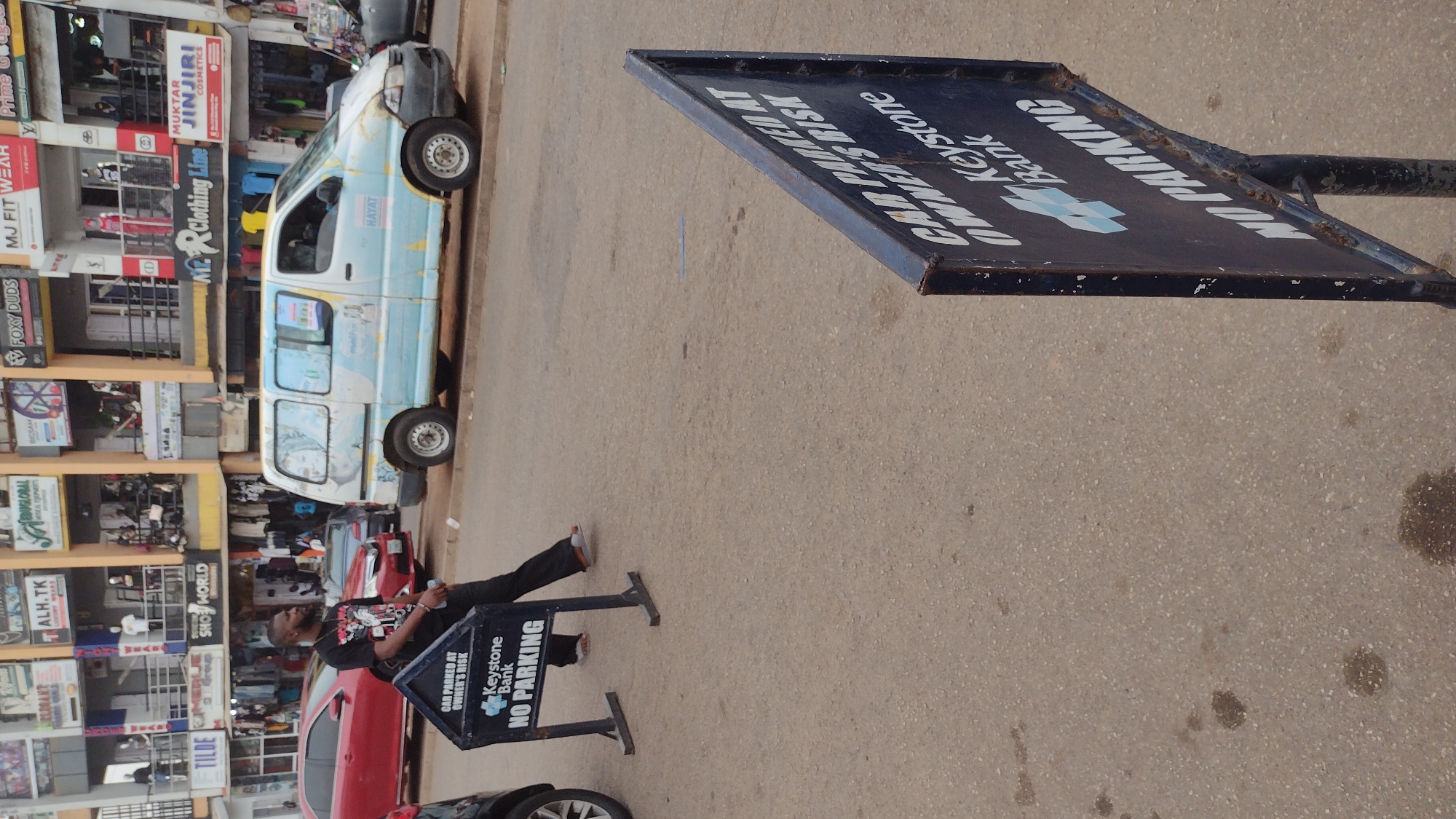 The image, taken sideways, shows a street scene in Jos, Nigeria. A man is sitting next to a "No Parking" sign for Keystone Bank, which also states "Car Parked at Owner's Risk." He is dressed in black, and next to the sign, a red car is parked. Further down the street, a white and blue vehicle is parked along a row of multi-story buildings. These buildings have shops with signs written in English, indicating various businesses such as clothing, cosmetics, and education services. The street itself is paved and shows signs of wear. The image is taken from a slightly elevated perspective.