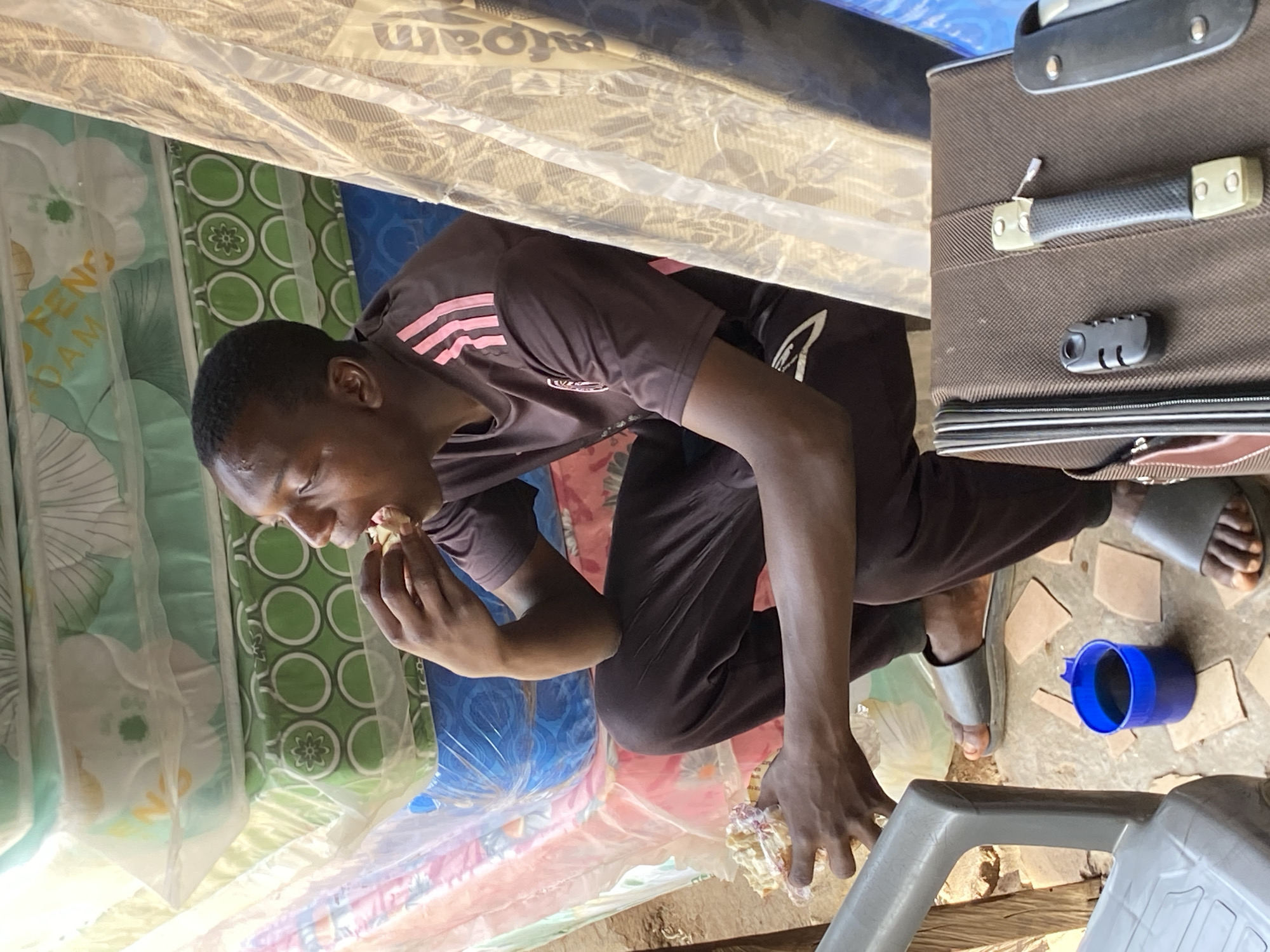 An individual is crouched, eating, inside a narrow space surrounded by mattresses wrapped in plastic. The mattresses display floral and geometric patterns. The person wears a brown t-shirt with pink stripes and dark-colored pants. He is positioned near a brown suitcase and wears grey sandals. Nearby is a blue plastic cup and a grey plastic container. The location is Bukuru, Nigeria.