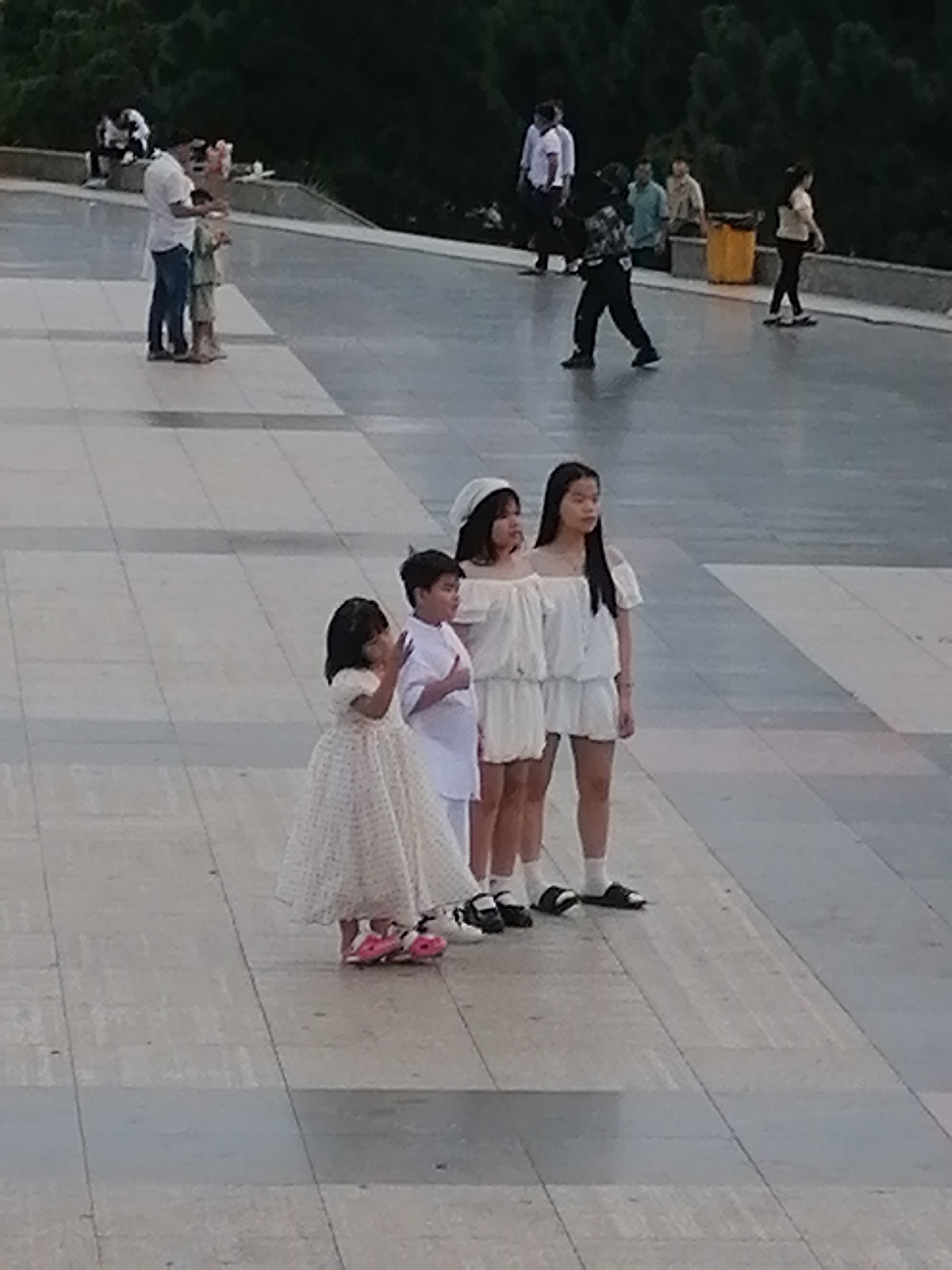 Four young people, three girls and one boy, stand in a line on a tiled plaza. The two leftmost figures are young girls. The leftmost girl wears a white dress with a patterned skirt and pink shoes with bows. The boy, to her right, wears a white shirt and pants. To the boy's right, a girl wears a white off-the-shoulder top and white shorts. The girl furthest to the right, also wearing a white off-the-shoulder top and white shorts, stands slightly behind the other three. They appear to be posing for a photograph. In the background, several adults are visible, some walking and others seated. Trees line the top of a sloping embankment behind the plaza. The scene is likely a public area in Da Lat, Vietnam, possibly a park or cultural site, given the attire of the individuals and the presence of trees.