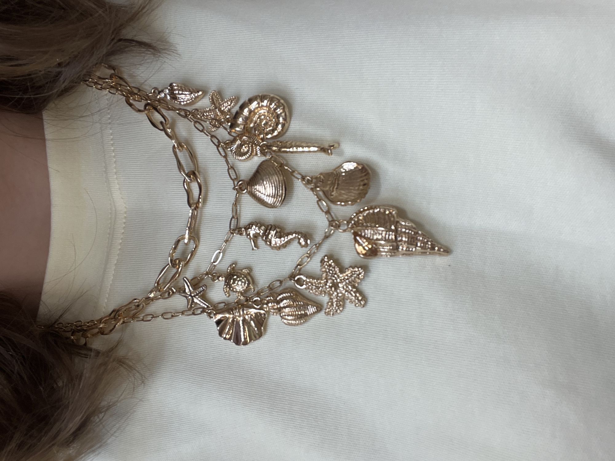 The image displays a close-up, slightly angled view of a multi-strand gold-toned necklace or bracelet adorned with numerous sea-themed charms. The accessory is worn by a person, whose neck and a section of light brown hair are visible on the left side of the frame.
The jewelry consists of several intertwined chains of varying thickness and link styles, from delicate links to more substantial oval-shaped ones. Attached to these chains are a collection of small, detailed charms, all in a matching gold finish. These charms primarily depict marine life and objects, including multiple starfish, various types of seashells (such as spiraled conch shells, ridged shells, and clam-like shells), a seahorse, and a small turtle.
The necklace is presented against a light-colored, possibly off-white or cream fabric, which appears to be part of the wearer's clothing, like a shirt or dress. The fabric exhibits a subtle horizontal texture or ribbing. The lighting is soft and even, accentuating the metallic luster of the charms and chains. The activity is limited to the display of the jewelry being worn. There are no notable location cues within the image itself, other than the general context provided of Podgorica, Montenegro. No text is visible in the image.