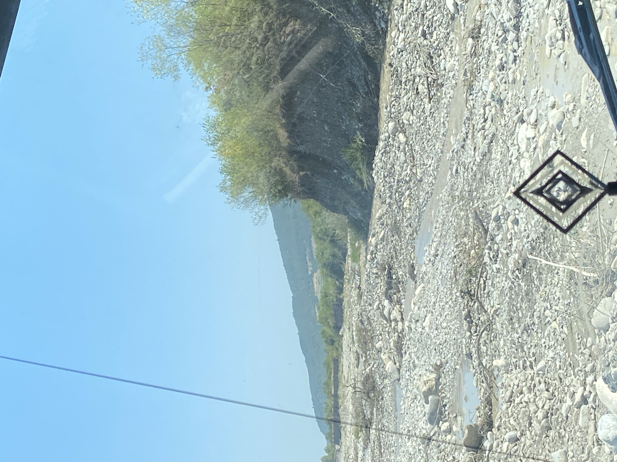 The image shows a dry riverbed filled with a multitude of rocks and debris, with sparse vegetation growing on the eroded banks. The sky is clear and blue, indicating a sunny day. A thin, dark line, possibly a wire or a reflection, stretches diagonally across the upper left portion of the image. In the foreground, a diamond-shaped ornament is visible, attached to the interior of a vehicle, with its reflection also appearing on the windshield. The landscape suggests a rural or mountainous area, consistent with the outskirts of Gorkhmazoba, Azerbaijan, where such riverbeds are common due to the region's topography and climate. No people are visible in the image.