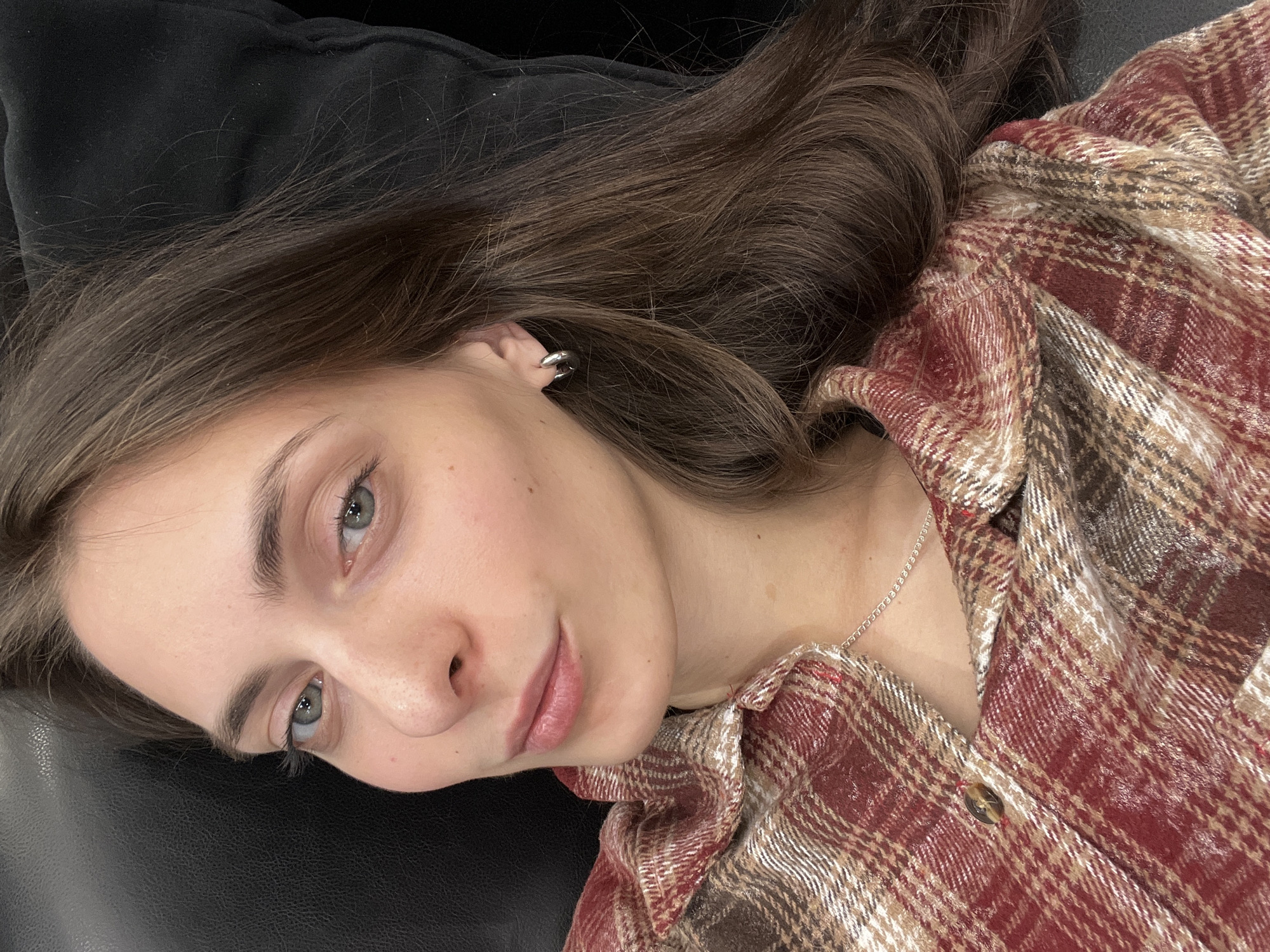A young woman with fair skin, light blue eyes, and brown hair is lying on a dark-colored surface. She is wearing a maroon and tan plaid shirt and a silver necklace. A silver earring is visible in her left ear. The background is a dark, solid color, possibly black leather. There is no evidence indicating the location is Krasnoyarsk, Russia, or any other specific location.