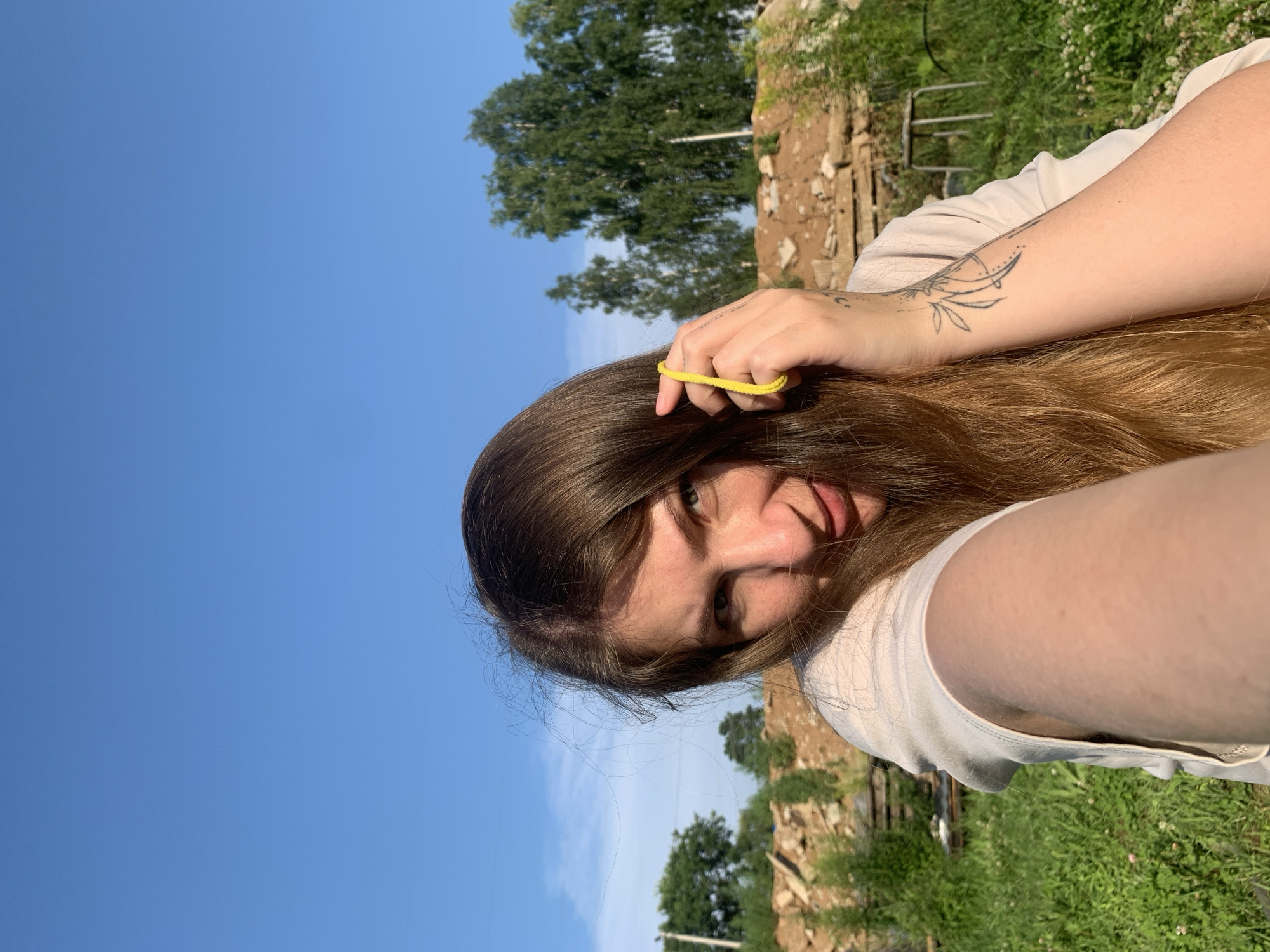 A young woman with long brown hair is taking a selfie outdoors. She is wearing a light-colored t-shirt and has a yellow elastic band in her hand, which she is holding near her hair. She has a tattoo on her left forearm. The background shows a bright blue sky with scattered clouds, green grass, and some piles of construction debris and trees. The lighting suggests it is daytime, likely late afternoon due to the warm glow. There is no visible indication of the specific location within Chaikovsky, Russia, beyond it being an outdoor, possibly semi-rural or developing area.