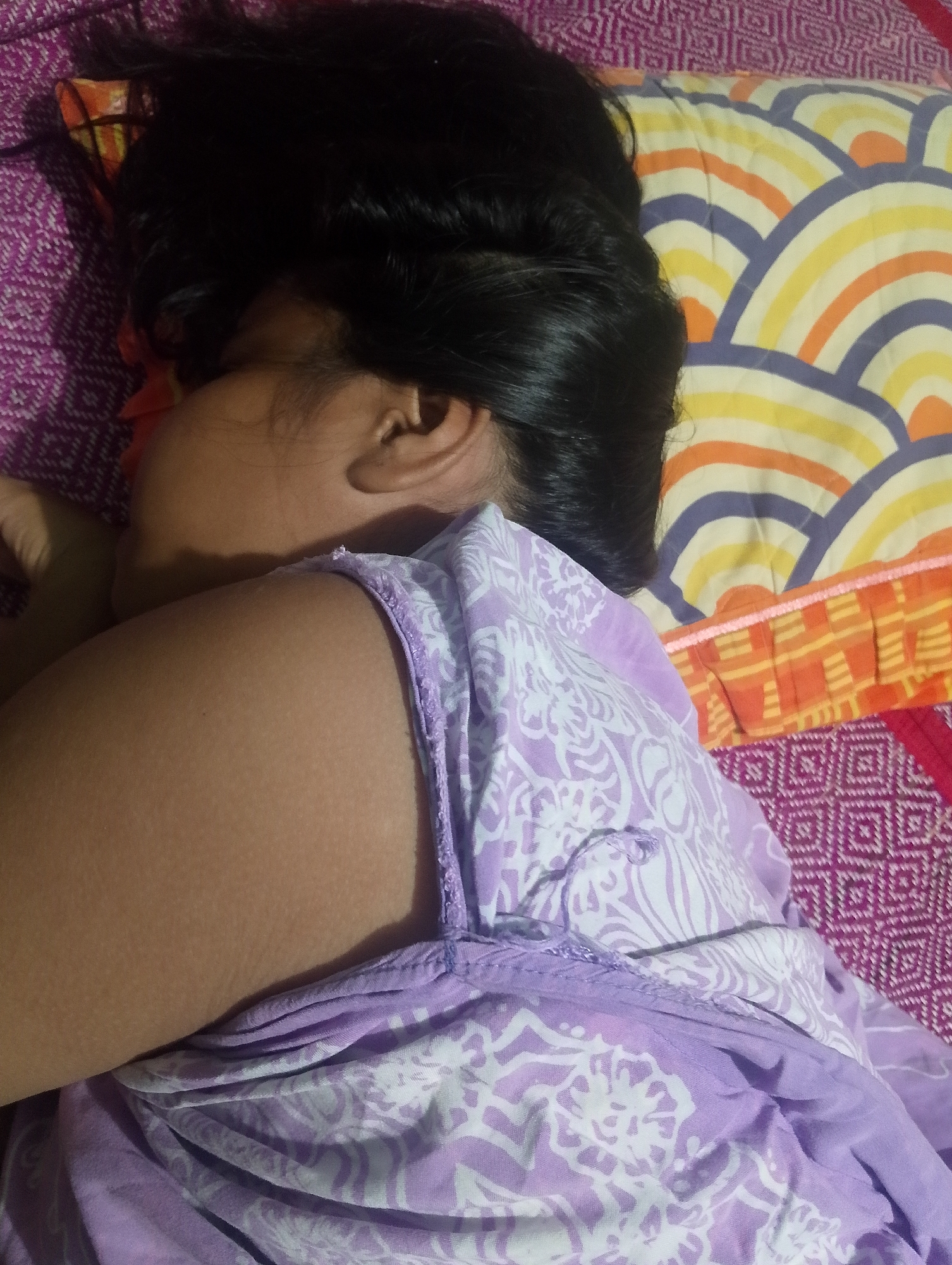 A person is sleeping on a bed in Serbelawan, Indonesia. The person is on their side, facing away from the camera. They have dark hair and are wearing a purple and white patterned shirt. The bed has a purple patterned sheet and a pillow with a colorful, abstract pattern. The lighting suggests it is nighttime or the room is dimly lit.