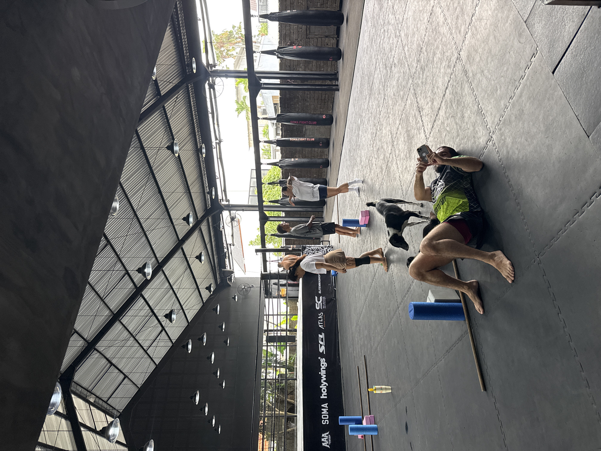 This image captures an indoor training session at a fight club or gym located in North Kuta, Indonesia. The setting is a spacious, industrial-style building with high ceilings featuring exposed metal trusses and corrugated roofing, along with dark grey interlocking puzzle mats covering the floor. A brick wall lines one side, from which several black cylindrical punching bags hang; two visible bags are branded with "SOMA FIGHT CLUB".
Approximately five individuals are present, engaged in various exercises. In the foreground, a woman in a white top and light shorts stands with her back to the viewer, possibly stretching. Further back, another person is performing a plank-like exercise. A bearded man lies on his side on the floor, holding a smartphone, seemingly documenting the activity. A black dog is also present, casually moving around the training area.
Scattered on the mats are various pieces of equipment, including blue and pink foam rollers, pink yoga blocks, and a wooden stick. Natural light illuminates the gym, and through the open structure, lush green foliage and palm trees typical of a tropical climate are visible, indicating it is daytime. A long black banner stretches along one wall, displaying text and logos such as "AAA", "TOWING", "SOMA", "holywings", "SFL", "ATLAS", "SC", and "ELITEMMASHOP". The scene portrays a functional and active training environment.