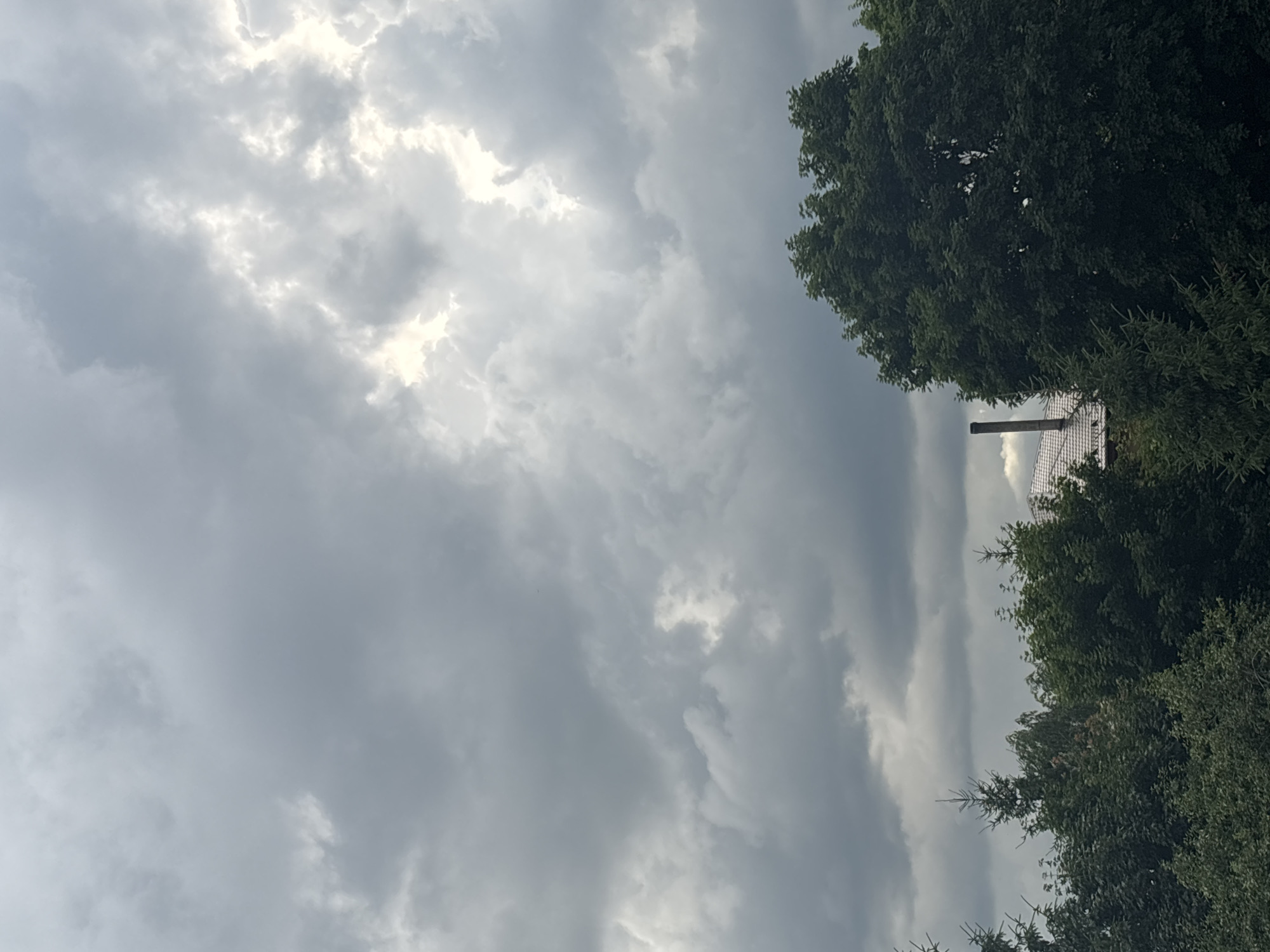 This is a vertical, high-angle shot of a cloudy sky with trees and a rooftop in the foreground. The sky is filled with a mixture of dark and light gray clouds, with patches of brighter sky visible through them, suggesting an overcast or possibly stormy weather condition.
On the right side of the image, a dense green canopy of trees dominates the lower portion, with their leaves appearing lush and full. Peeking out from behind the trees, a tiled rooftop of a building is visible, along with a dark, cylindrical chimney. The trees are likely deciduous, given their full foliage.
The scene appears to be set outdoors, likely in a residential or semi-rural area given the presence of the rooftop and chimney amidst the greenery. The lighting suggests it is daytime, but the heavy cloud cover creates a muted, diffused light. There is no visible text in the image. The overall mood conveyed is somewhat somber due to the cloudy sky and dark foliage. The specific location is identified as Ostrov, Russia.