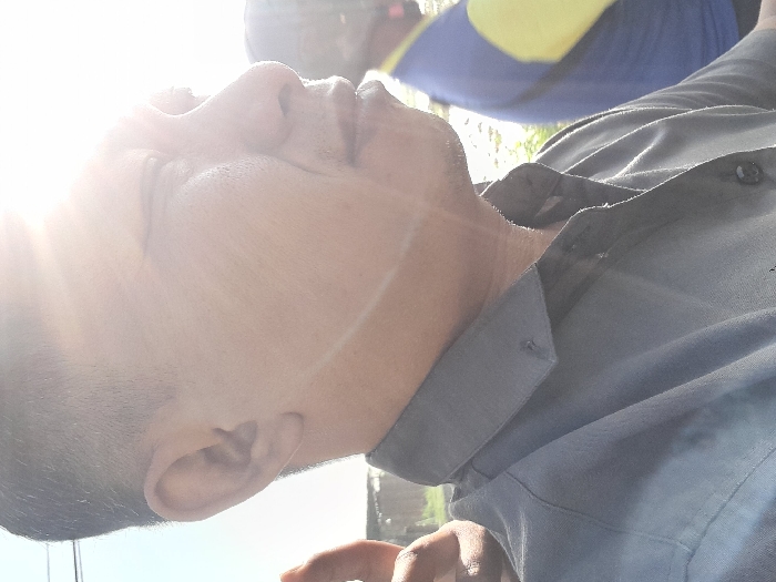 A man's face is illuminated by sunlight. He's wearing a grey button-down shirt. Another person is partially visible in the background, wearing a blue and yellow shirt. The shot is close-up, focusing on the man's profile and the side of his head.