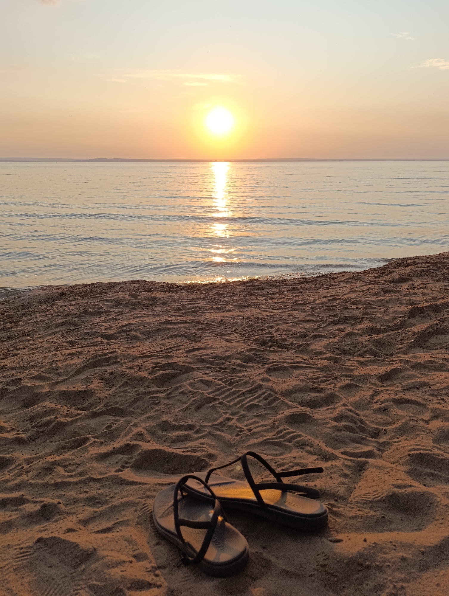 In the foreground, a pair of black flip-flops rests on textured sandy ground, their straps casting shadows. The sand shows imprints, possibly from footprints. Beyond the sand, a calm body of water stretches to the horizon, reflecting the warm glow of a setting sun. The sun, a bright orb, is positioned in the upper center of the frame, its light creating a shimmering path across the water. The sky above transitions from a pale yellow near the horizon to a light blue at the top. The overall scene evokes a peaceful, tranquil atmosphere.