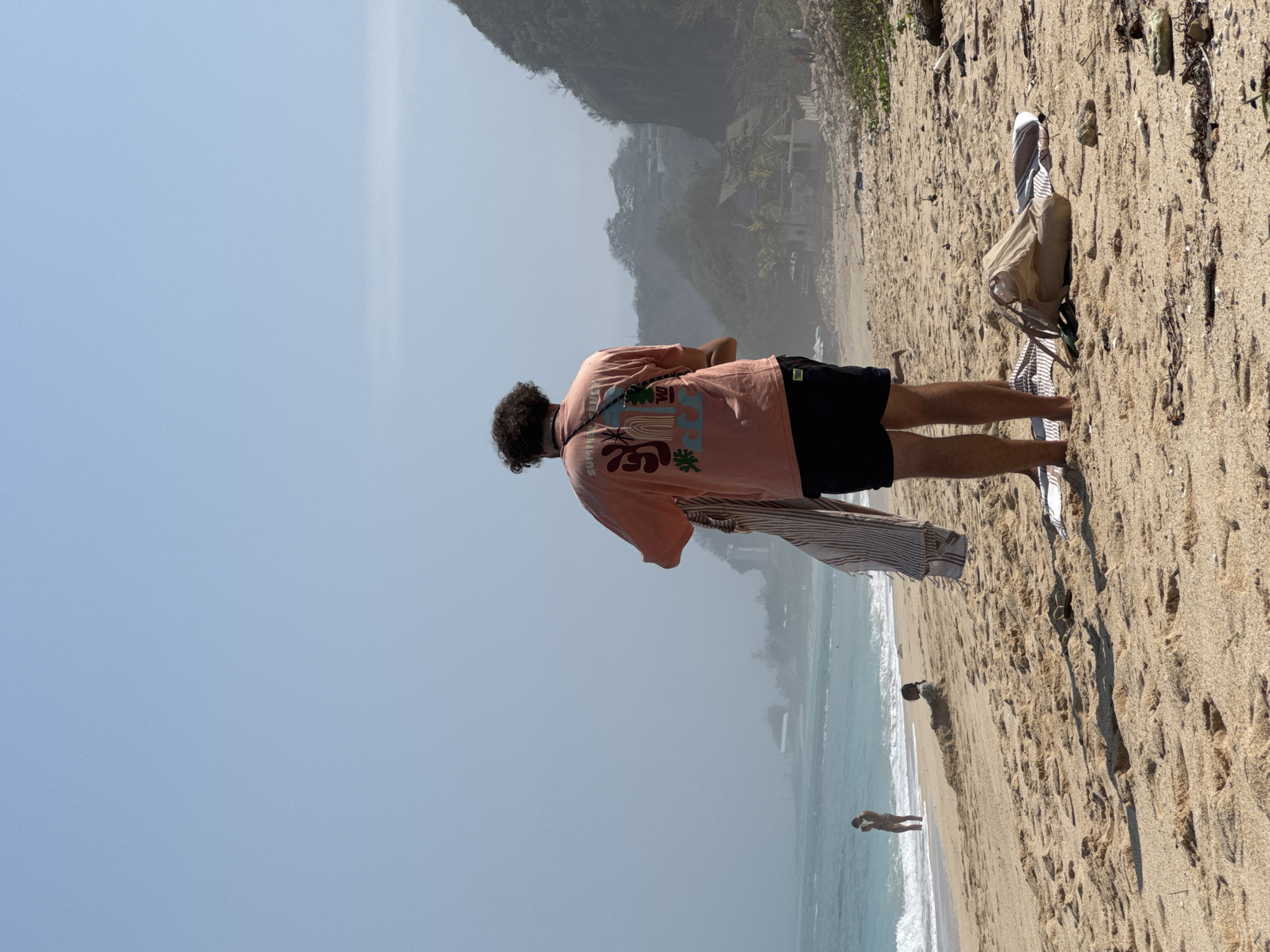 The image depicts a beach scene in Kuta Selatan, Indonesia. In the foreground, a person is standing with their back to the camera, wearing a coral-colored t-shirt with a graphic design and dark shorts. They are holding a striped towel. A beige beach bag and a striped towel are on the sand to their right. In the middle ground, a person is sitting partially buried in the sand, facing left. Further in the distance, a person is standing in the turquoise water near the shore. The background shows a hazy coastline with vegetation and buildings on a cliffside, with the ocean stretching out towards the horizon under a clear sky. The beach is sandy, with some debris like dried seaweed and twigs scattered on the surface.