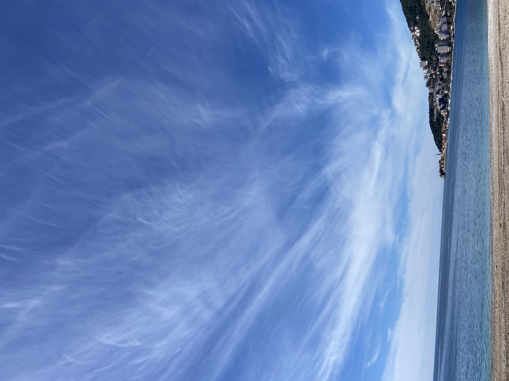 The image depicts a bright, daytime scene along the coast in Budva, Montenegro. The sky is a vibrant blue with streaks of cirrus clouds stretching across the majority of the frame. To the right, a narrow strip of the Adriatic Sea is visible, with gentle waves near the shore. Adjacent to the sea, a pebble beach extends, marked by a light tan color. Further inland, a dense cluster of buildings is nestled along a hillside, indicative of the urban landscape of Budva. The buildings appear to be residential and commercial structures, typical of a coastal city. There are no people visible in the image. The perspective is likely from a ground-level position looking upwards and towards the coastline.