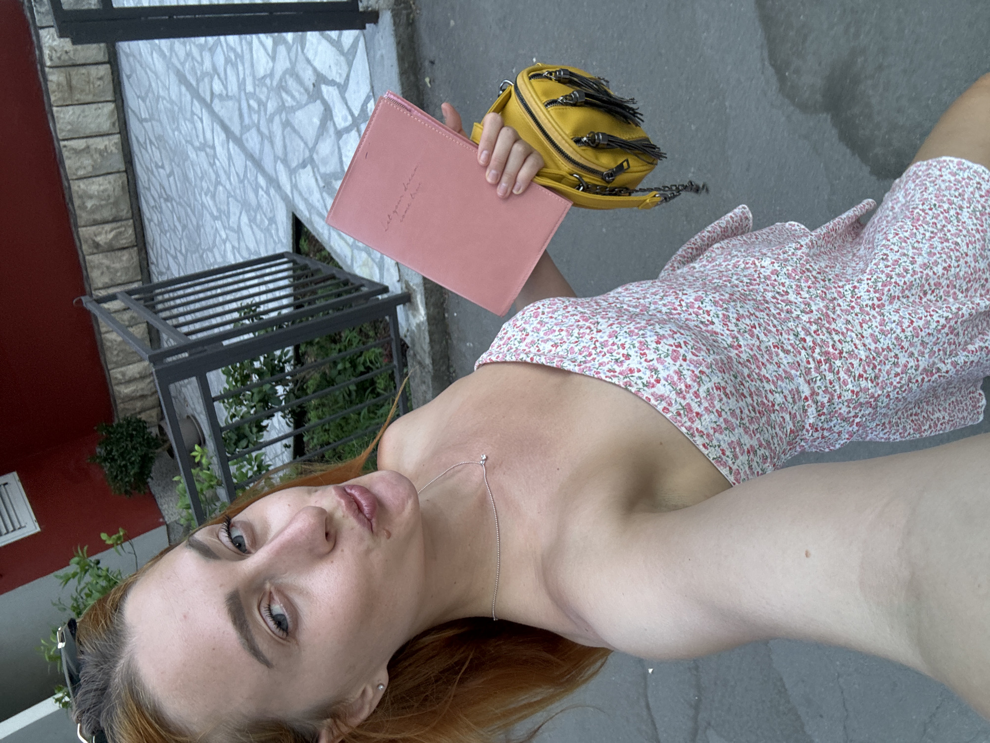 A woman with red hair and blue eyes, wearing a strapless floral dress and a silver necklace, takes a selfie outdoors. She is holding a pink notebook and a yellow purse with tassels. The background shows a stone-paved area and a metal fence. The image appears to be taken in Budva, Montenegro, based on the architectural style and paving.