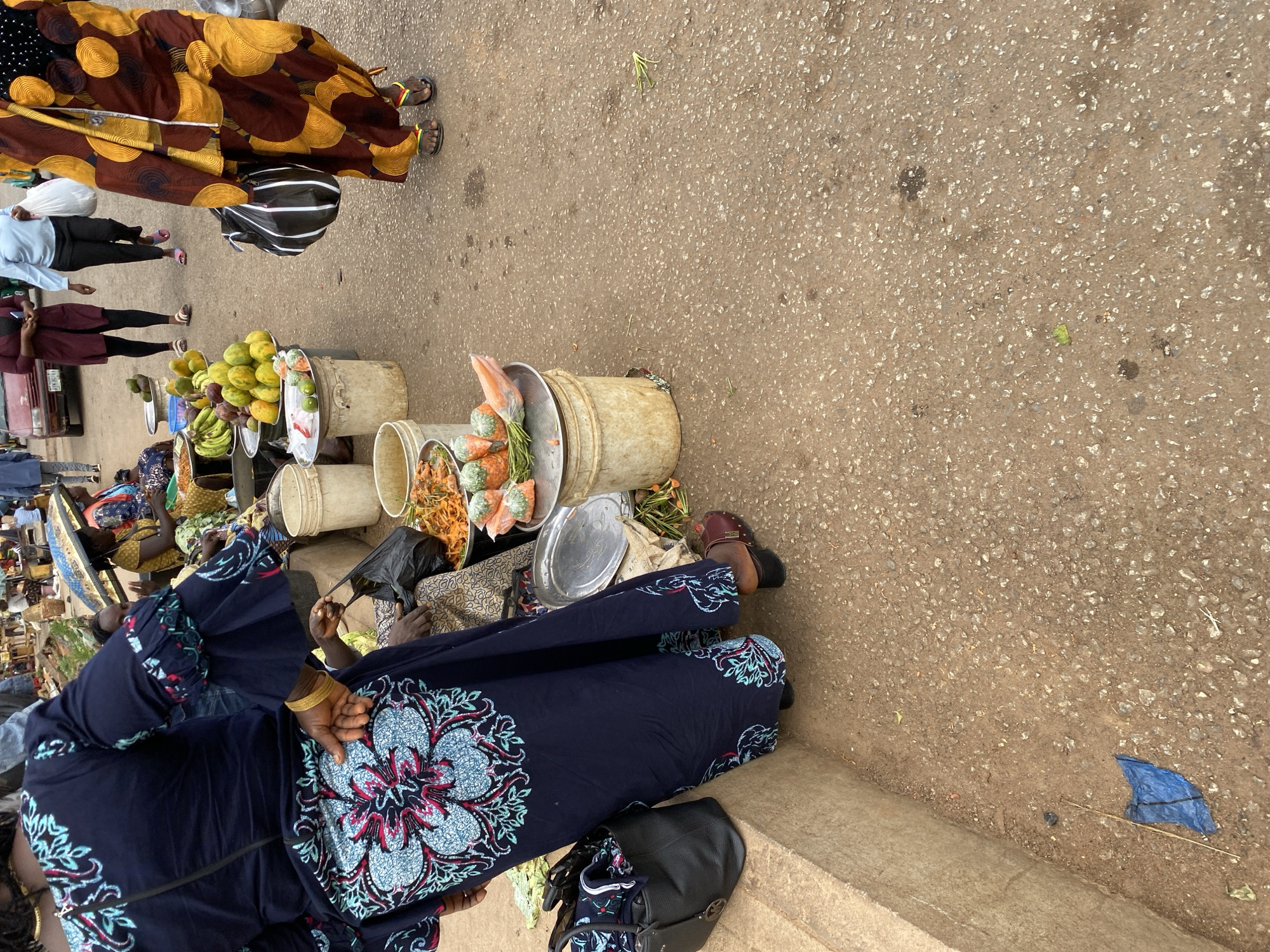 The image depicts a bustling street market scene in Bukuru, Nigeria. Numerous vendors are selling their goods, including fruits (mangoes, bananas, papayas) and snacks, displayed in buckets and trays. People are walking through the market. Several vendors are seated near their wares. One is wearing a patterned blue dress and gold bangles. A blue plastic sheet lies on the ground.